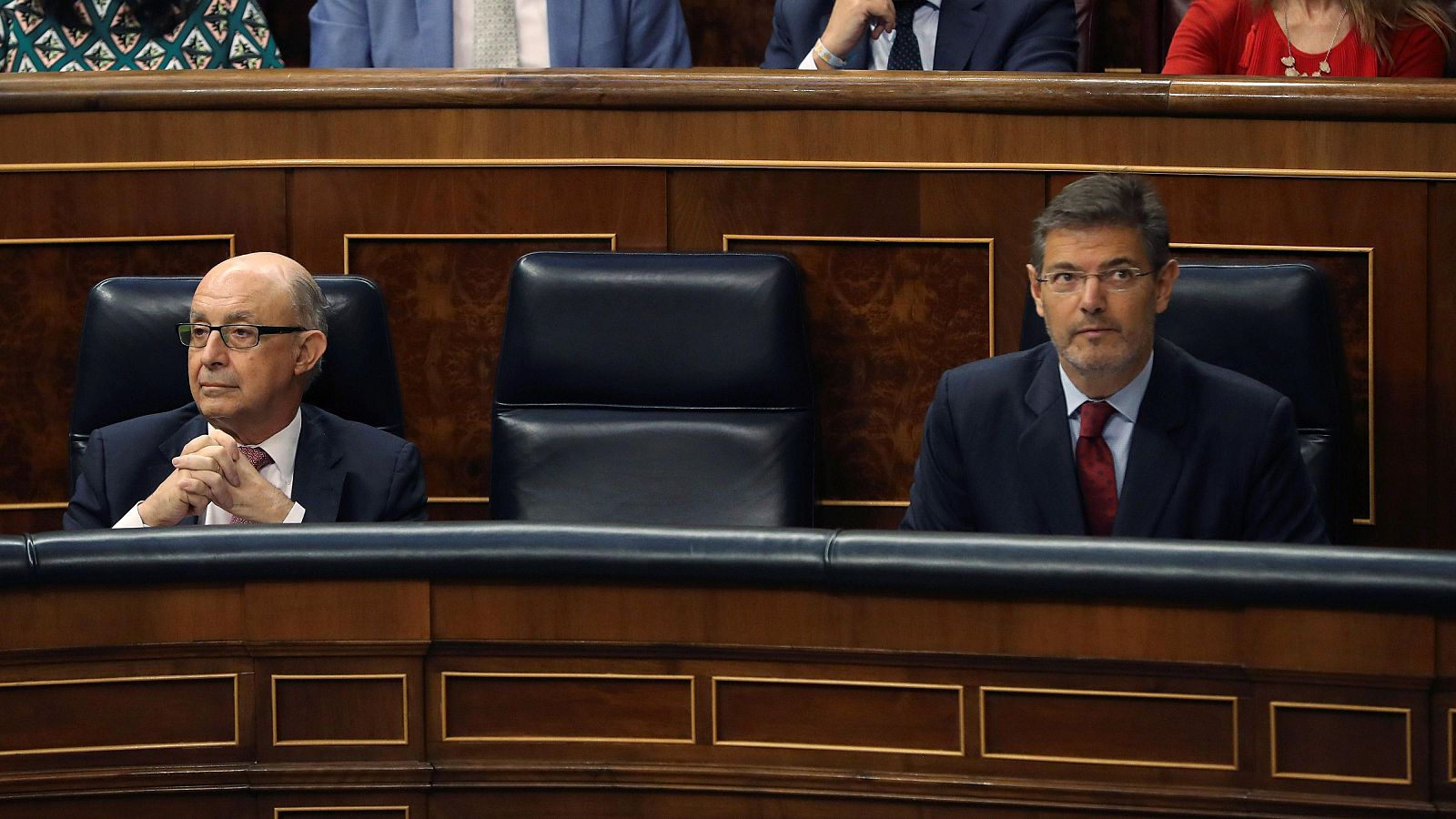 Dos hombres en traje oscuro y corbatas de colores diferentes sentados en asientos de cuero en un hemiciclo.  Se observa parte de la bancada y otras personas en el fondo.