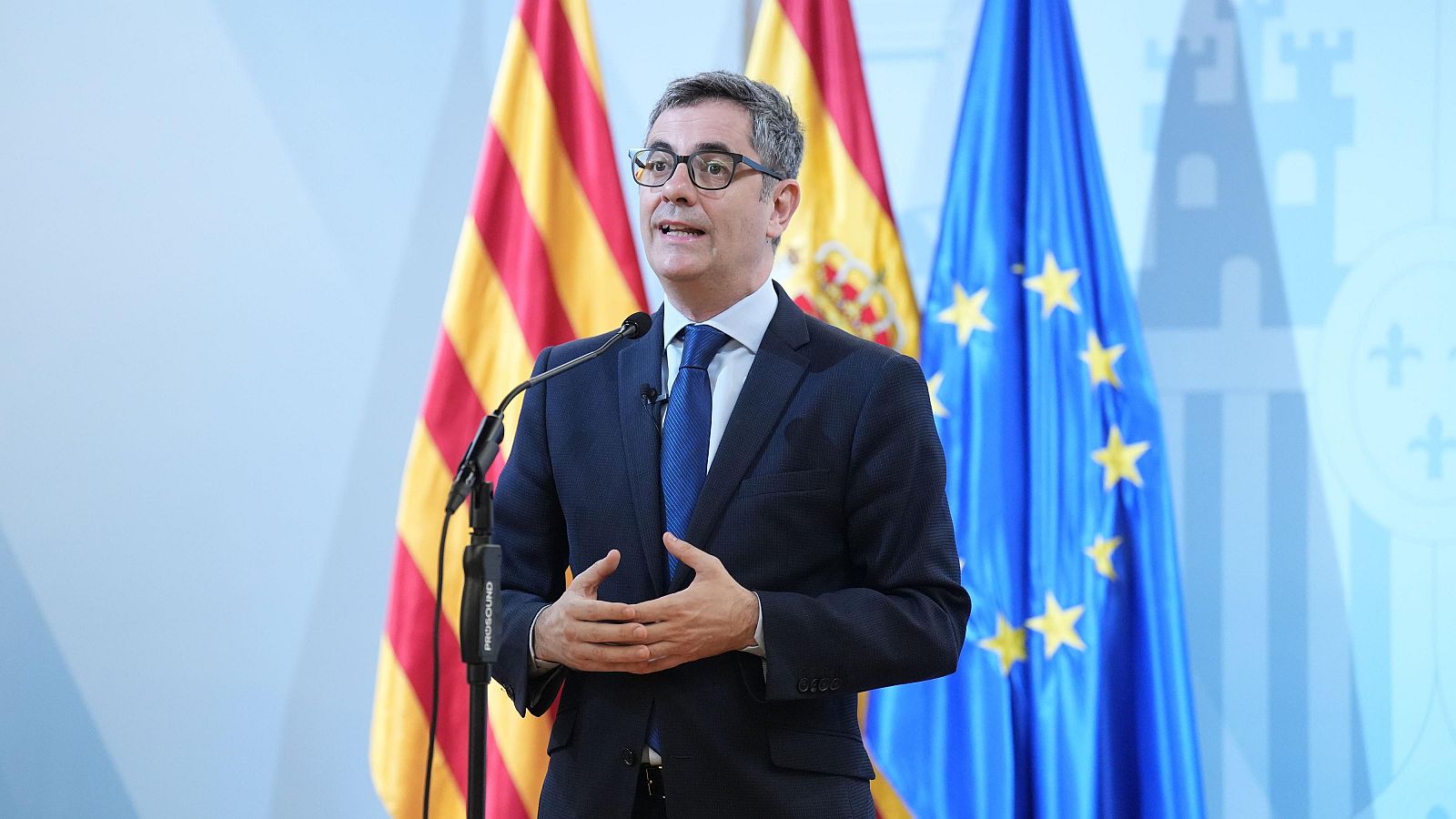 Comparecencia de un ministro en traje oscuro y corbata azul, de pie ante micrófonos y un fondo con banderas españolas y europeas.