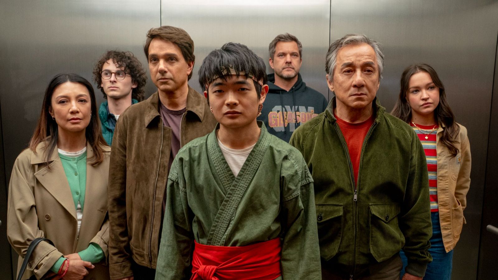 Siete personas en un ascensor, un joven con gi verde oliva y cinturón rojo destaca entre los demás con expresiones serias.