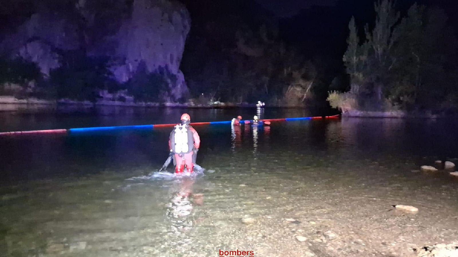 Búsqueda nocturna de desaparecidos en río. Bomberos con trajes de buceo trabajan en aguas poco profundas usando una barrera flotante. Escena dramática.