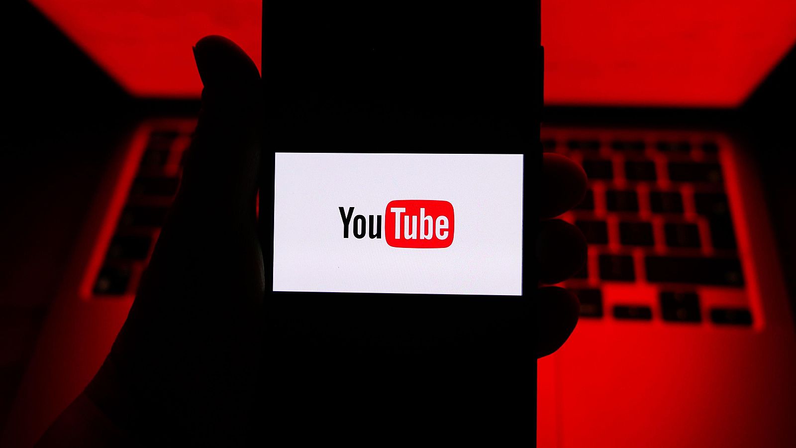 Mano sujetando móvil con el logo de YouTube; fondo con teclado y luz roja.  Imagen relacionada con nuevas normas sobre directos de menores.