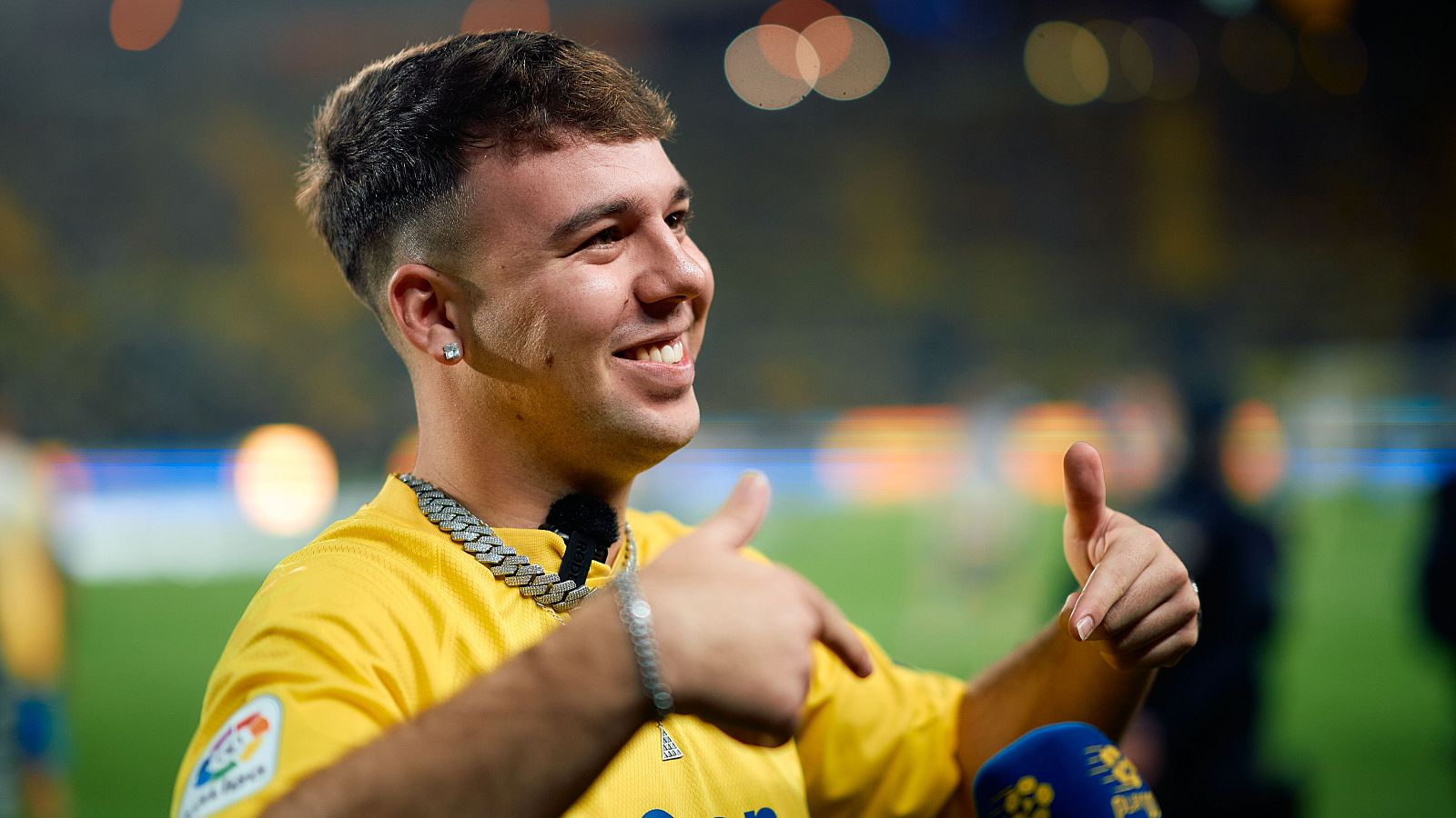 Cantante con camiseta amarilla y cadena, gesto positivo con pulgares arriba, micrófono visible, posible concierto en estadio.