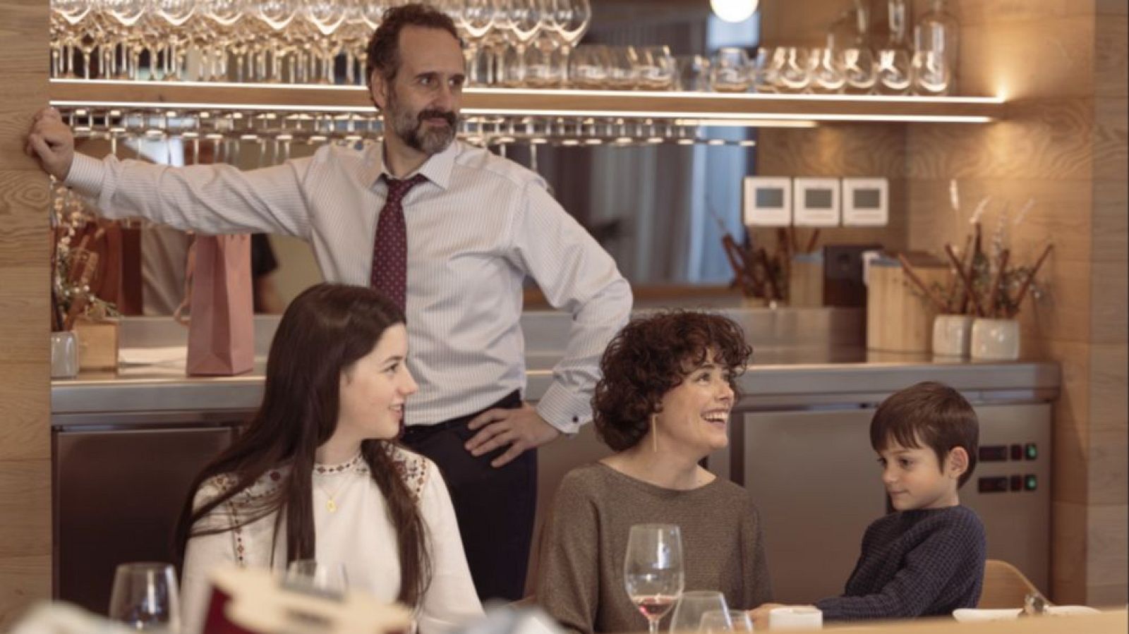 Familia en restaurante: padre de pie, madre e hijos sentados a la mesa con copas de vino. Ambiente tranquilo.
