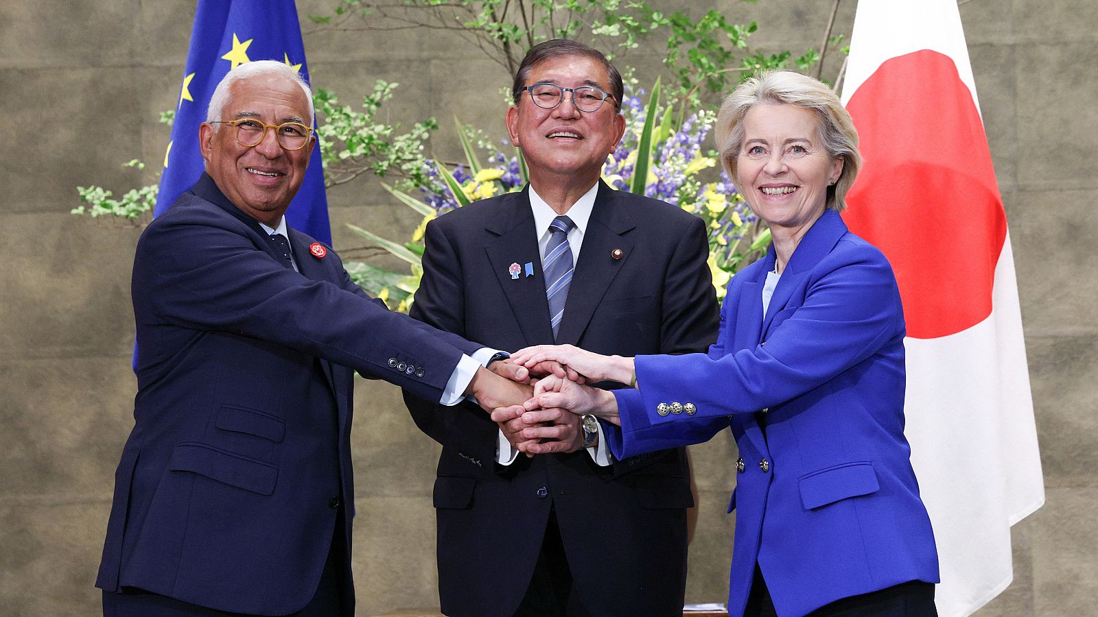 Tres líderes internacionales, con las manos juntas, muestran cooperación entre la UE y Japón.  Banderas de ambos países presentes en el fondo.