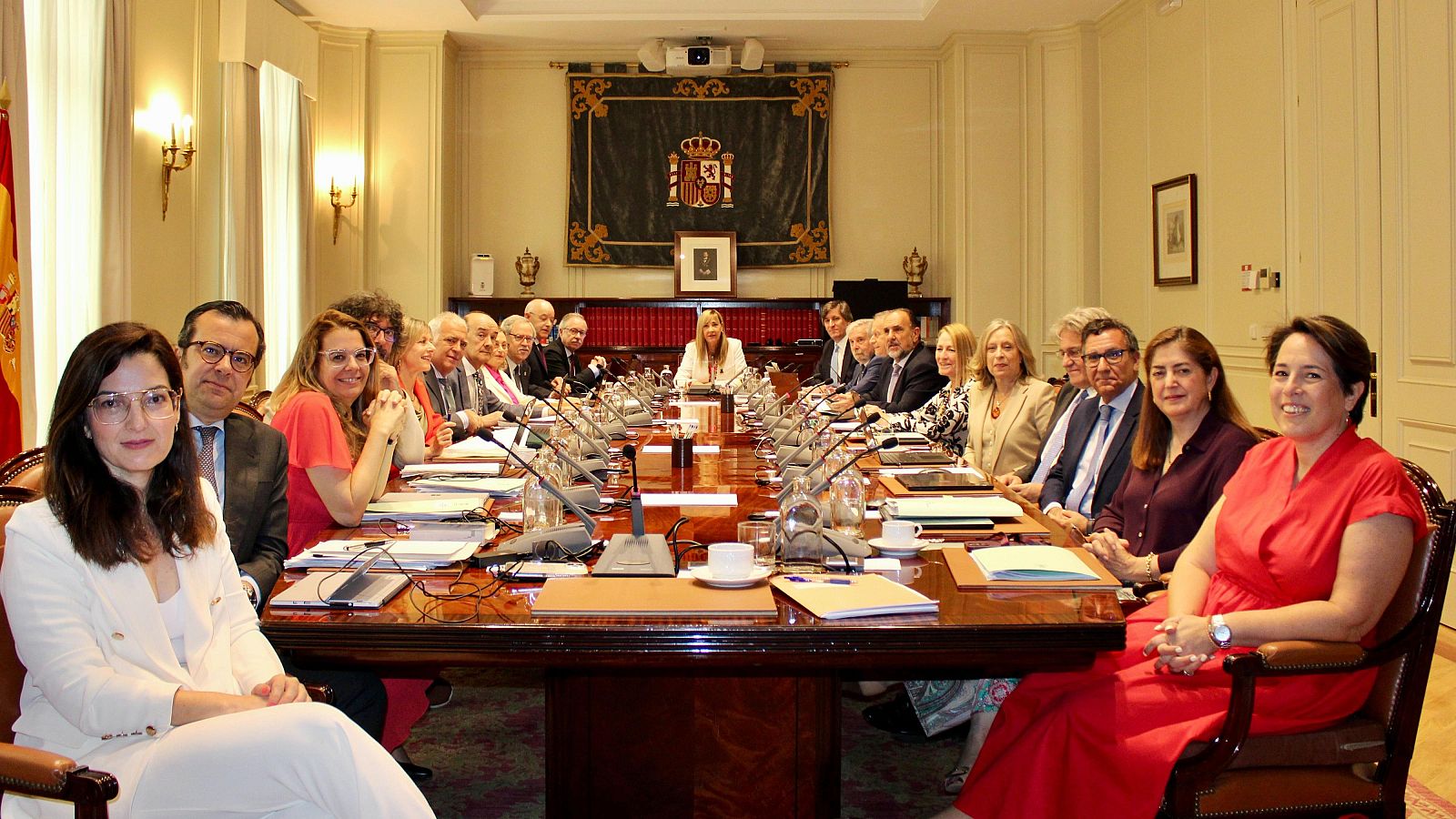 Reunión formal en una sala con una larga mesa de madera, micrófonos y carpetas.  Se observa la bandera de España en la pared.