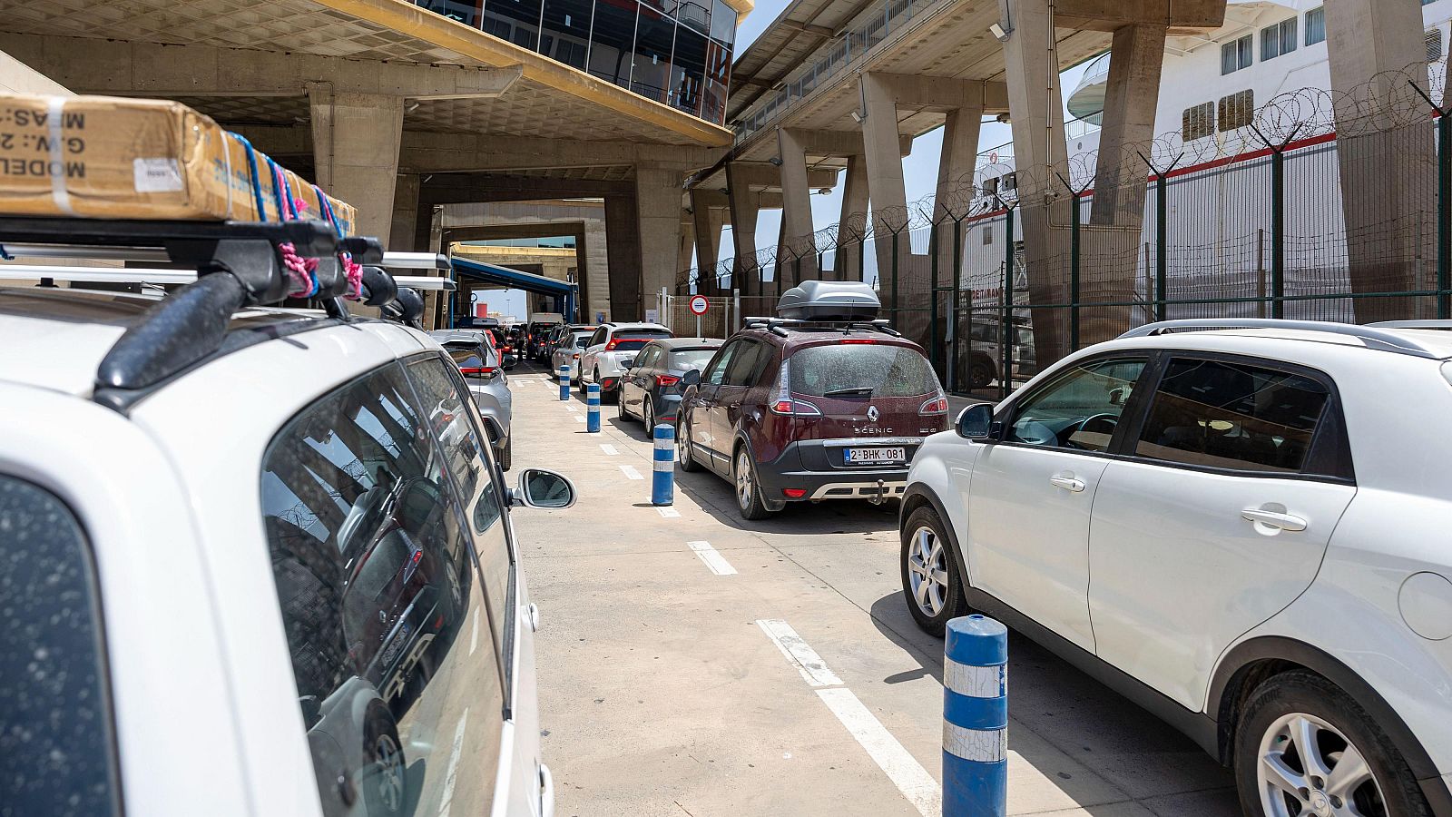 Coches con equipaje esperan en fila para embarcar en un ferry, delimitada por bolardos.  Se observa parte de un ferry y una valla al fondo.