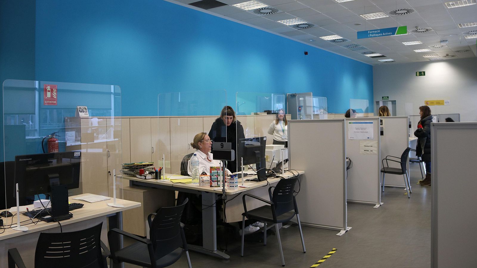 Oficina con puestos de trabajo, ordenadores y personal.  Mamparas y extintor presentes. Iluminación empotrada y señalización de emergencia.