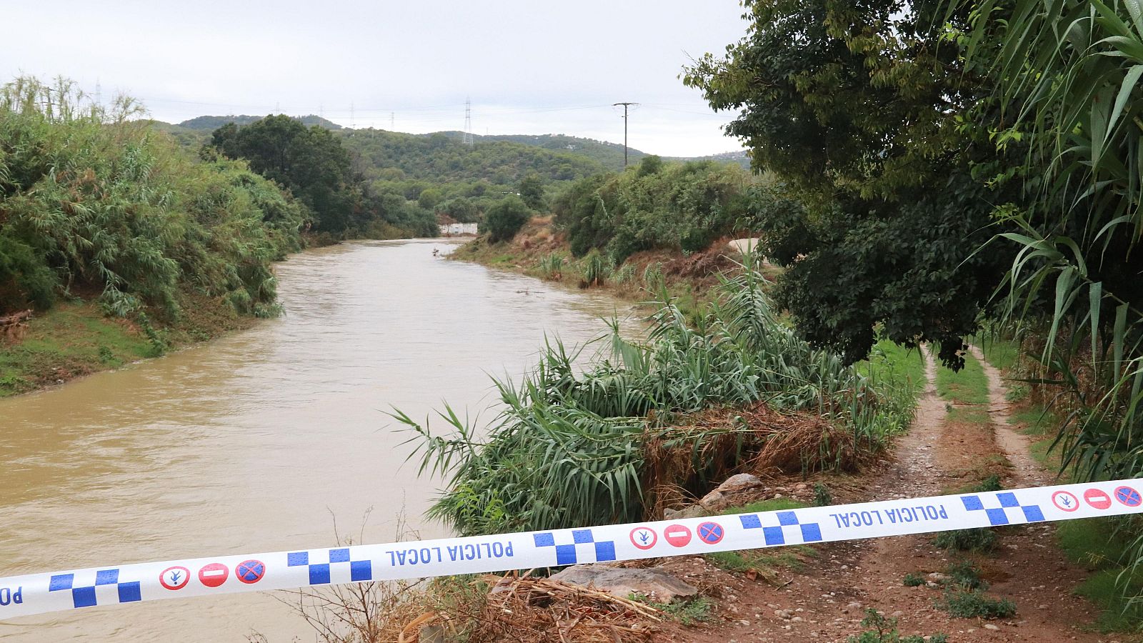 Río crecido con aguas turbias y camino bloqueado por cinta de Policía Local. Vegetación abundante y colinas al fondo.