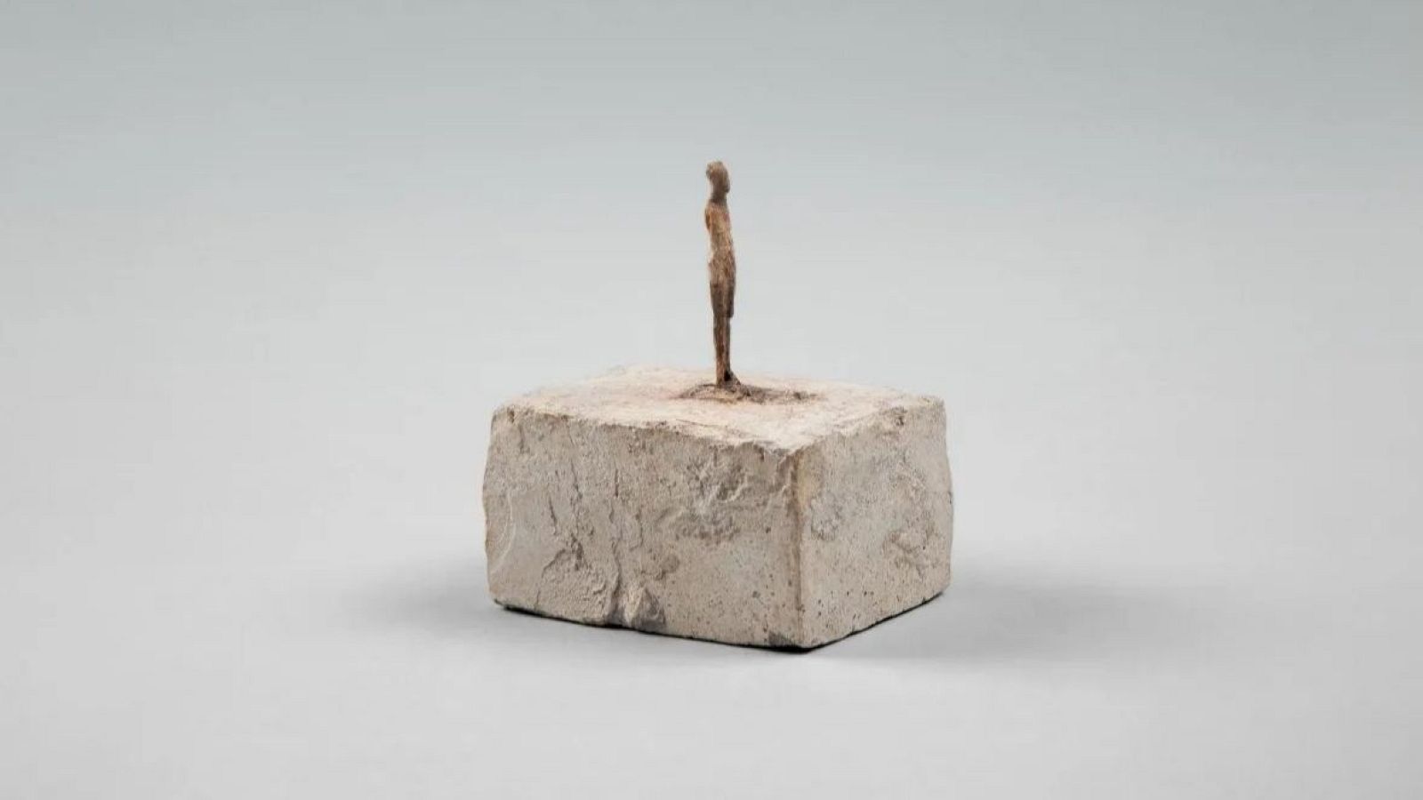 Escultura estilizada marrón oscuro, figura delgada y alargada sobre pedestal rectangular gris claro de textura rugosa. Fondo gris uniforme.