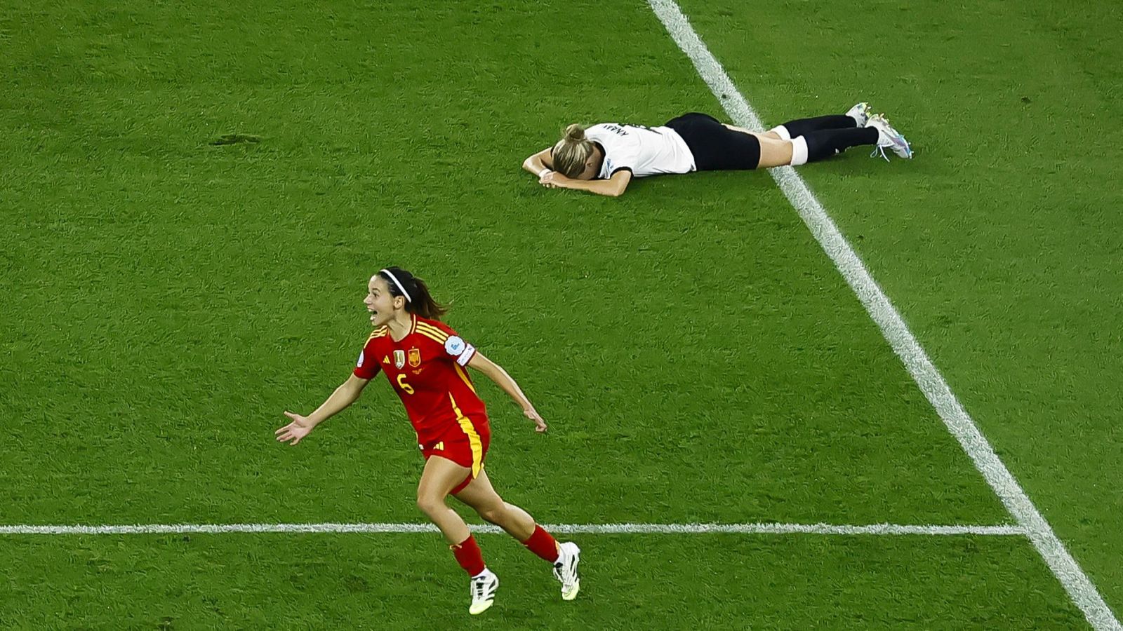 Jugadora con camiseta roja y número 6 celebra un gol, mientras otra, de blanco, yace en el césped mostrando frustración.  Momento de alta tensión en un partido de fútbol.