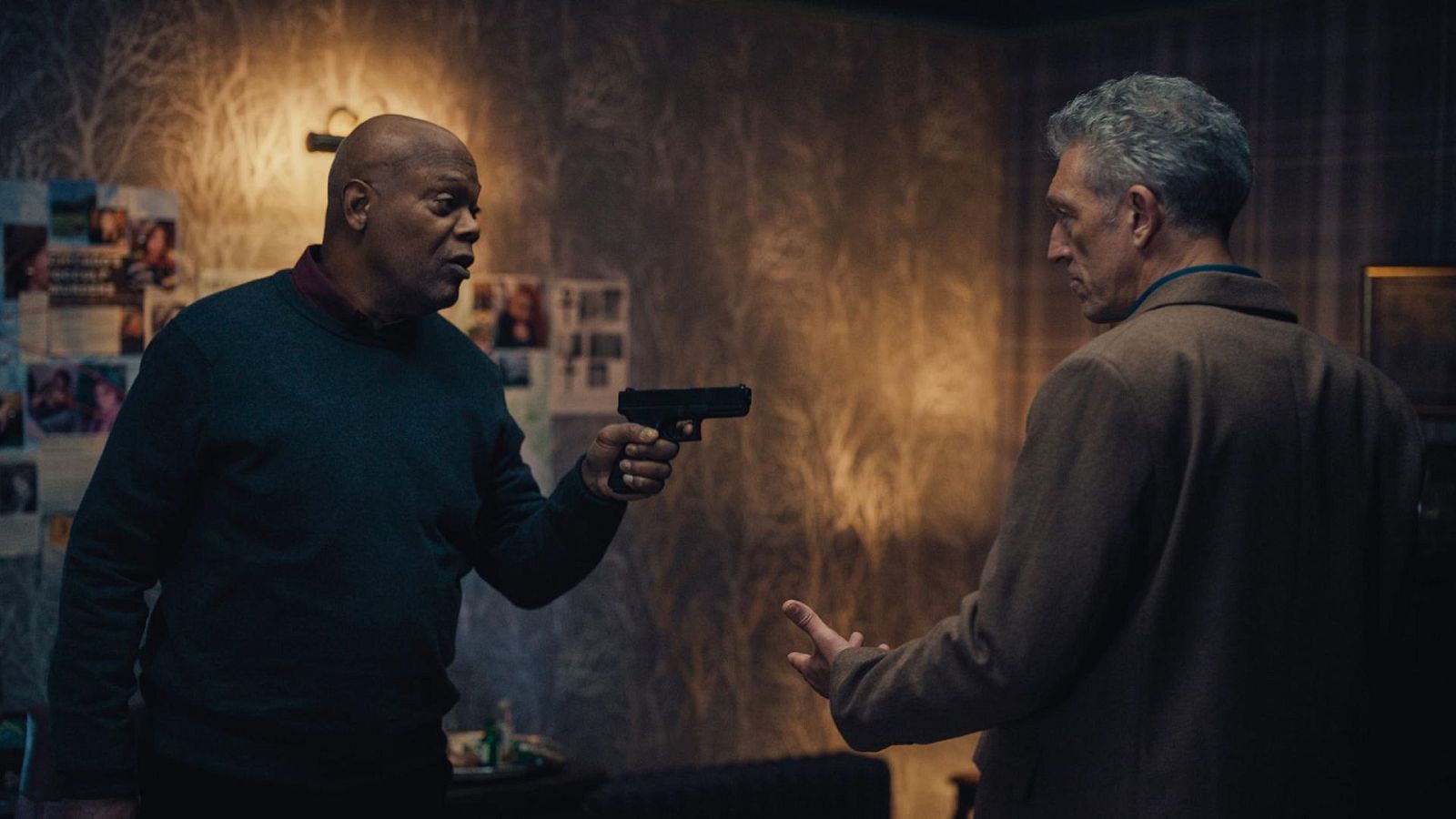 Samuel L. Jackson apunta con una pistola a otro hombre en una habitación con fotos en la pared; la escena transmite tensión.