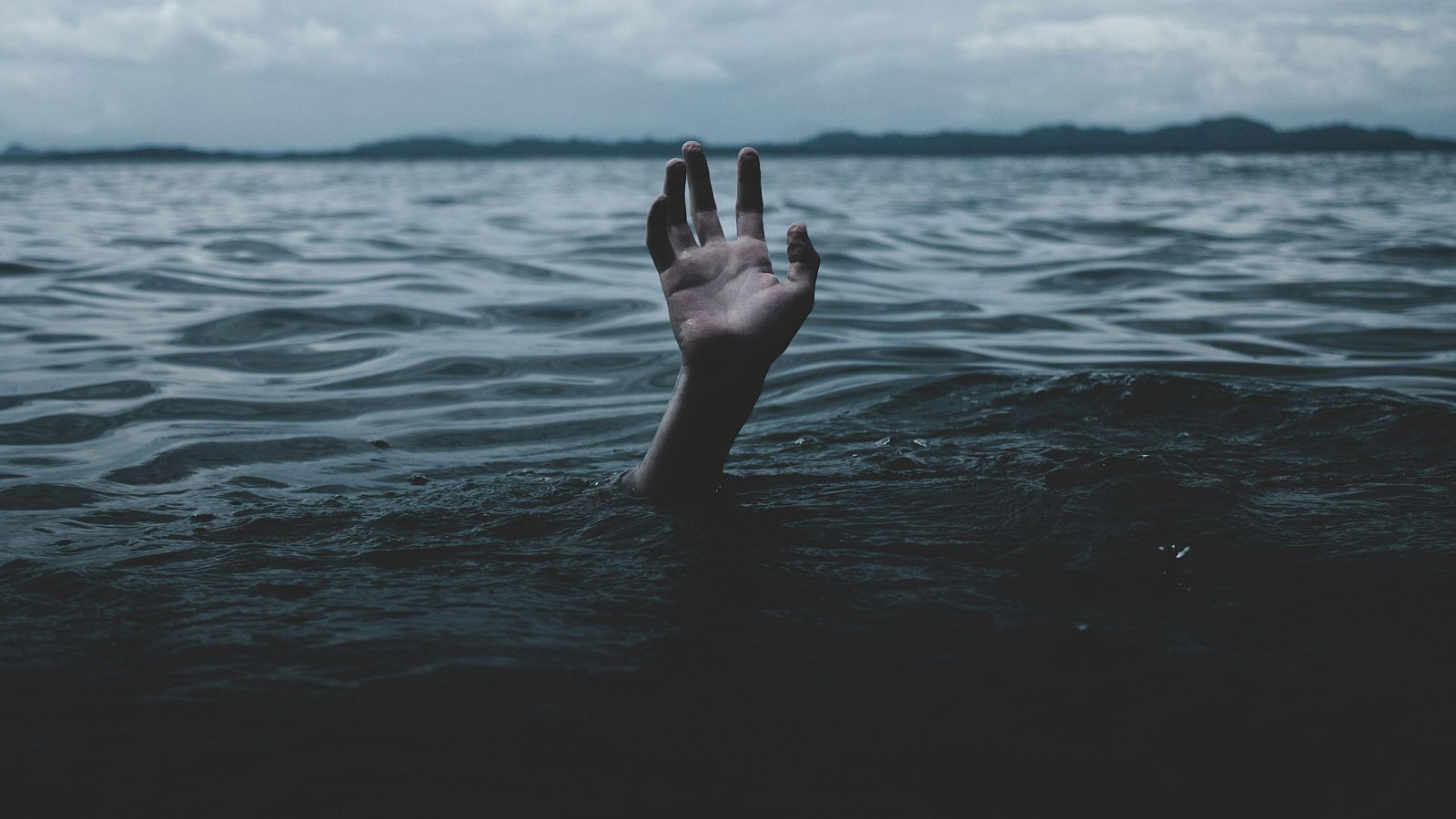 Mano emergiendo de agua oscura y con dedos extendidos; cielo nublado y fondo montañoso. Atmósfera sombría.