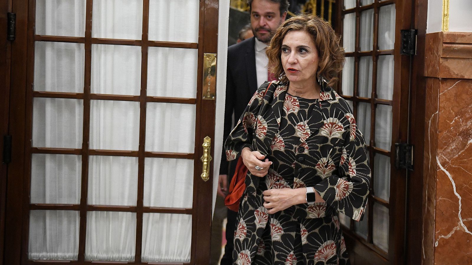 La ministra de Hacienda, María Jesús Montero, viste un abrigo oscuro estampado y bolso rojo, junto a una puerta de madera con cristales. Al fondo, un hombre con traje.