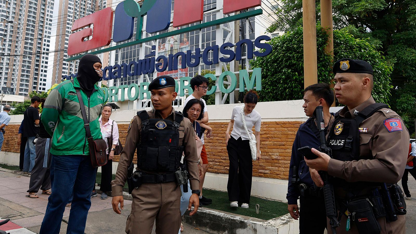 Escena posterior a un tiroteo: dos policías, uno con móvil, atienden la situación junto a civiles conmocionados. Un hombre con chaqueta verde y pasamontañas está presente.