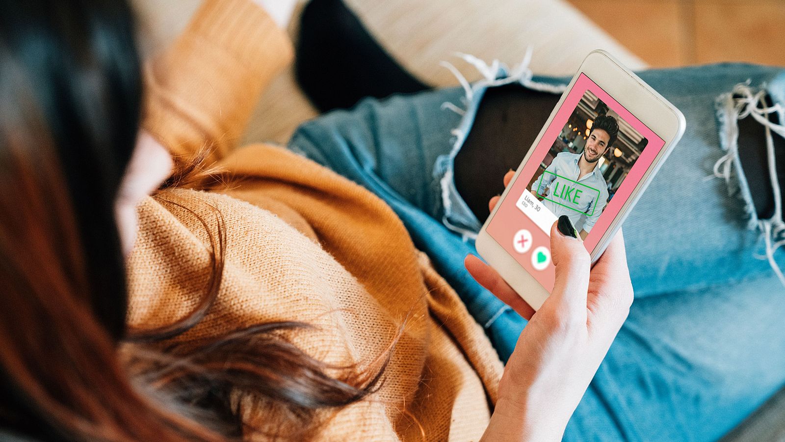 Mujer usando app de citas; su dedo está sobre el botón de "me gusta" (corazón verde) en un perfil masculino. Interfaz sencilla con opciones de aceptar o rechazar.