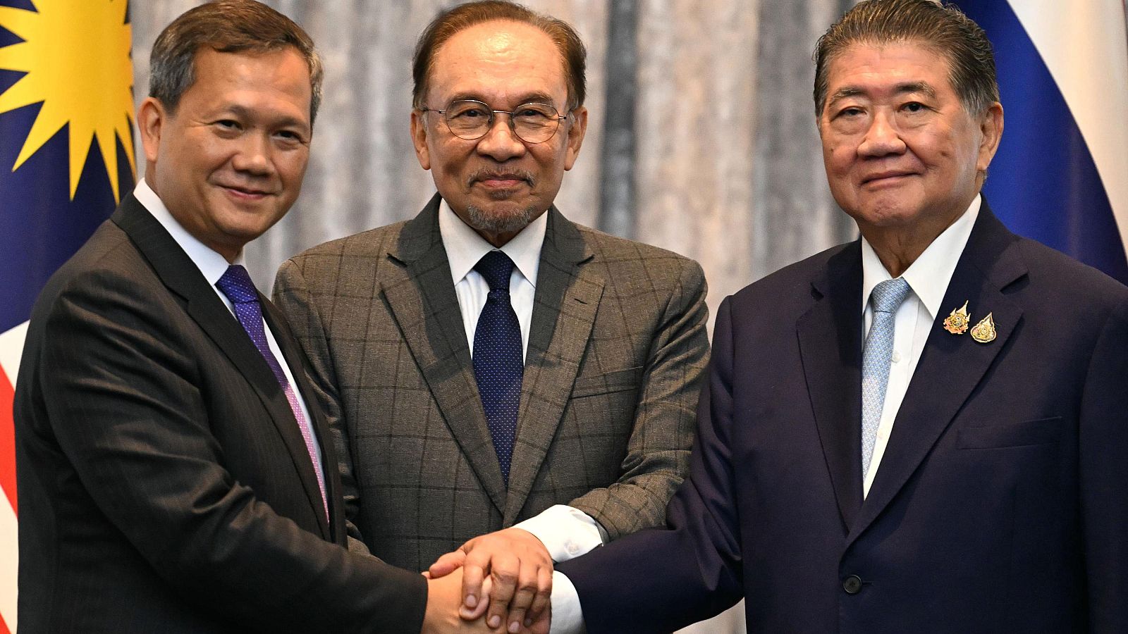 Acuerdo político: tres hombres en traje estrechan sus manos ante banderas de Malasia y Tailandia. Posible mediación.