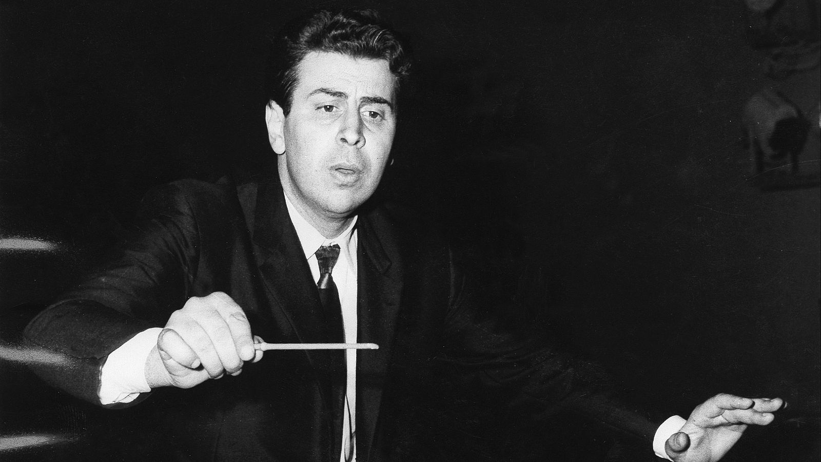 Mikis Theodorakis, dirigiendo una orquesta; viste traje oscuro y corbata, usando una batuta. Imagen en blanco y negro, fondo desenfocado.