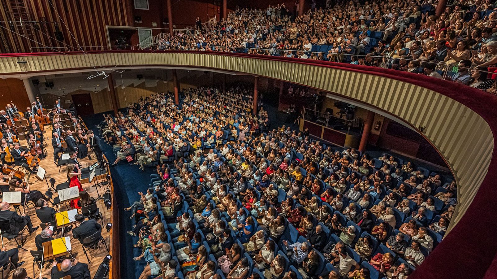 Auditorio lleno durante un concierto de orquesta y coro.  Músicos en escena con diversos instrumentos, público atento y aplaudiendo.