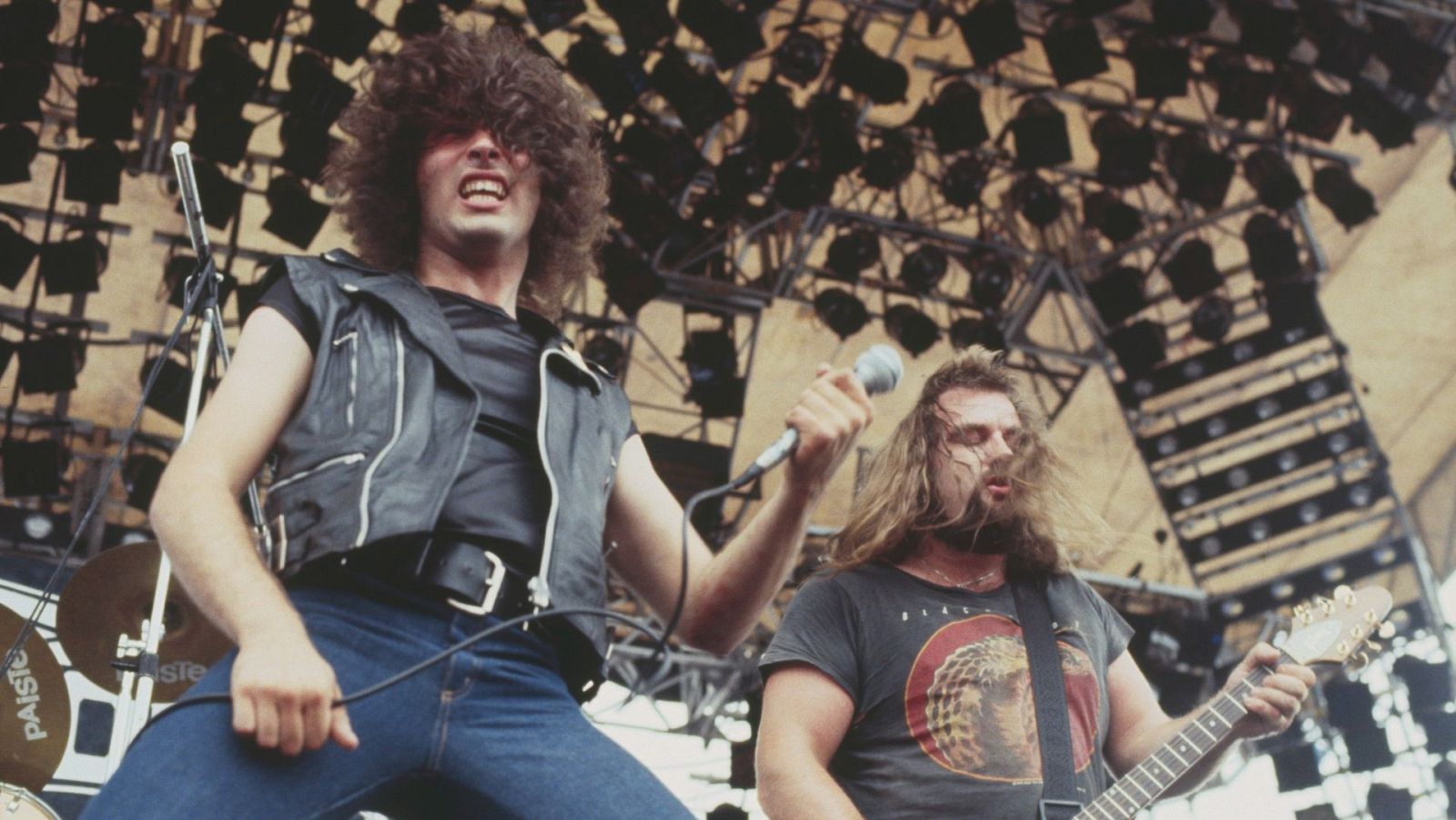 Concierto al aire libre en 1981: vocalista con micrófono y pelo largo, guitarrista con guitarra eléctrica y pelo largo. Escenario metálico con focos visibles.