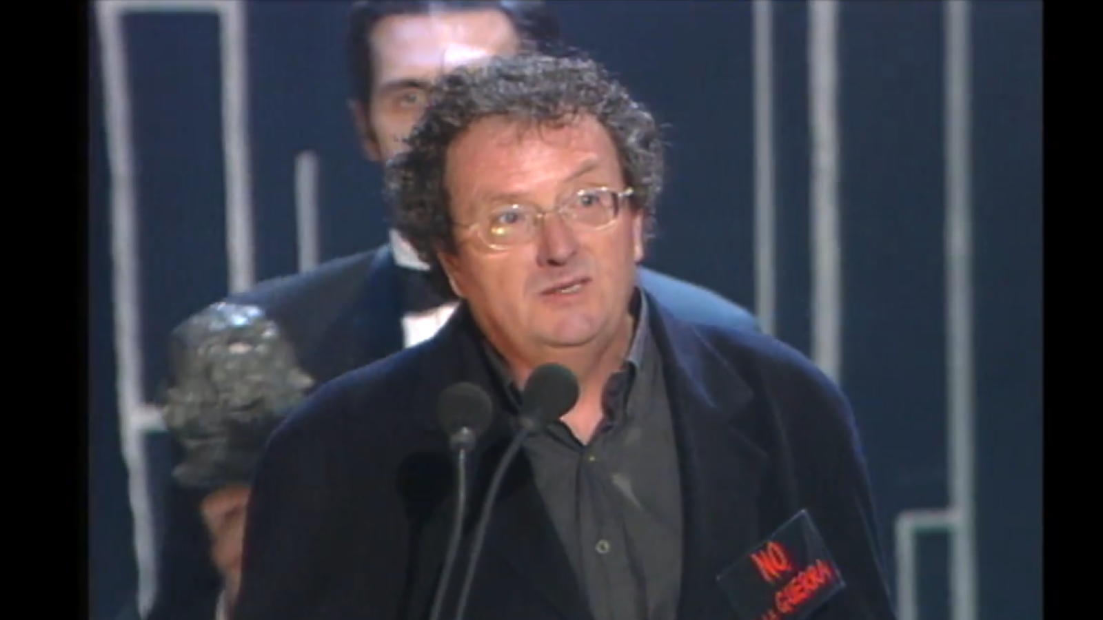 El cineasta Pere Joan Ventura agradece un premio en una gala, con chaqueta oscura, gafas y una pegatina 'NO a la guerra' en la solapa.