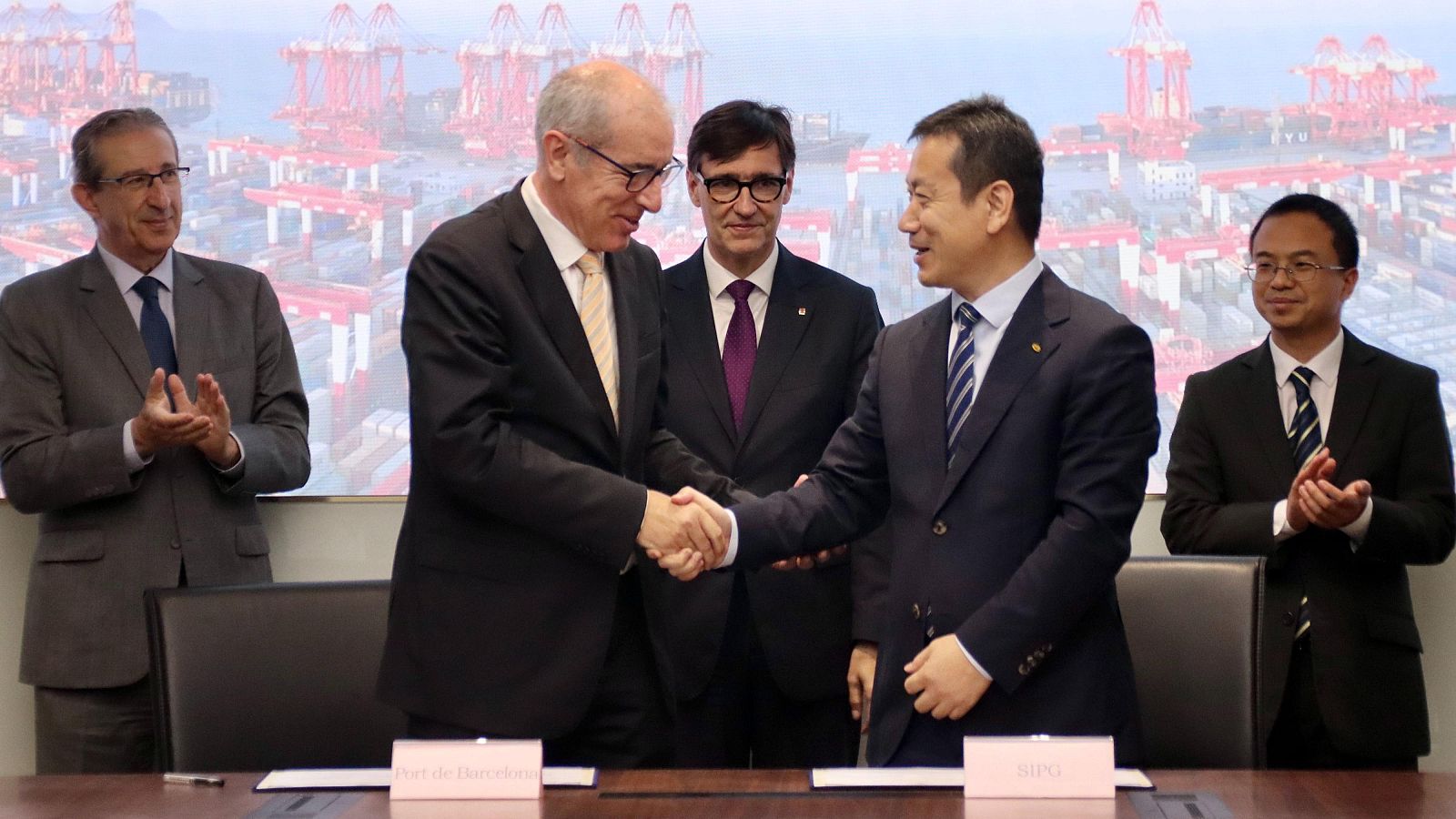 Convenio entre el Port de Barcelona y el Port de Shanghai.  Sus presidentes se dan la mano, rodeados de asistentes en traje que aplauden.  Ambiente formal.