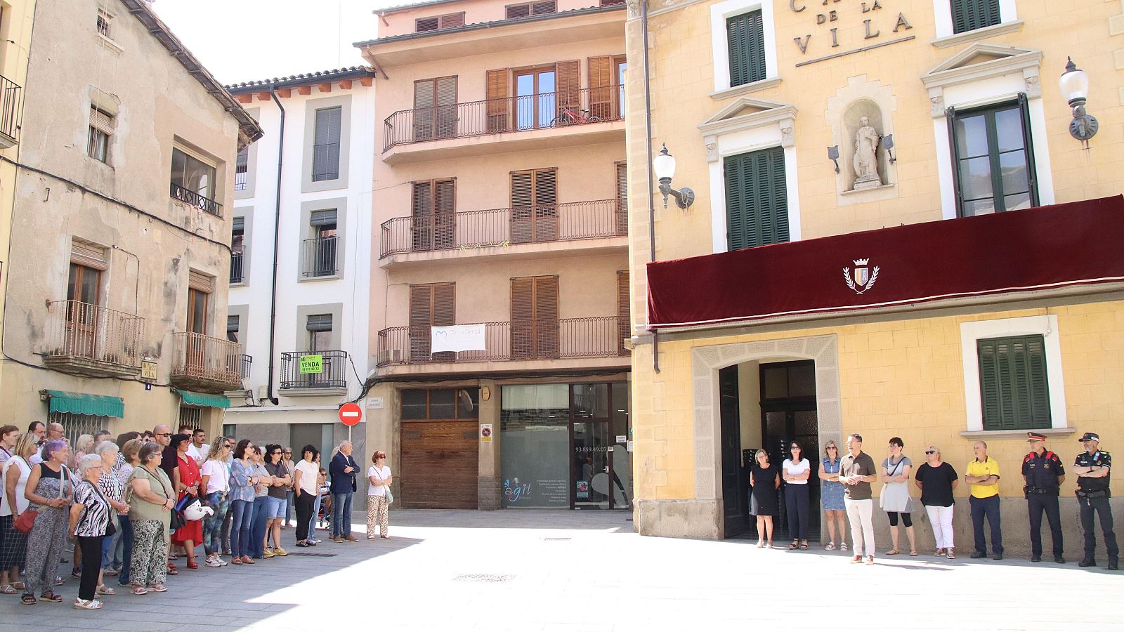 Minuto de silencio en una plaza, frente a un edificio con una pancarta.  Personas, mayoritariamente a un lado, muestran respeto y solemnidad.