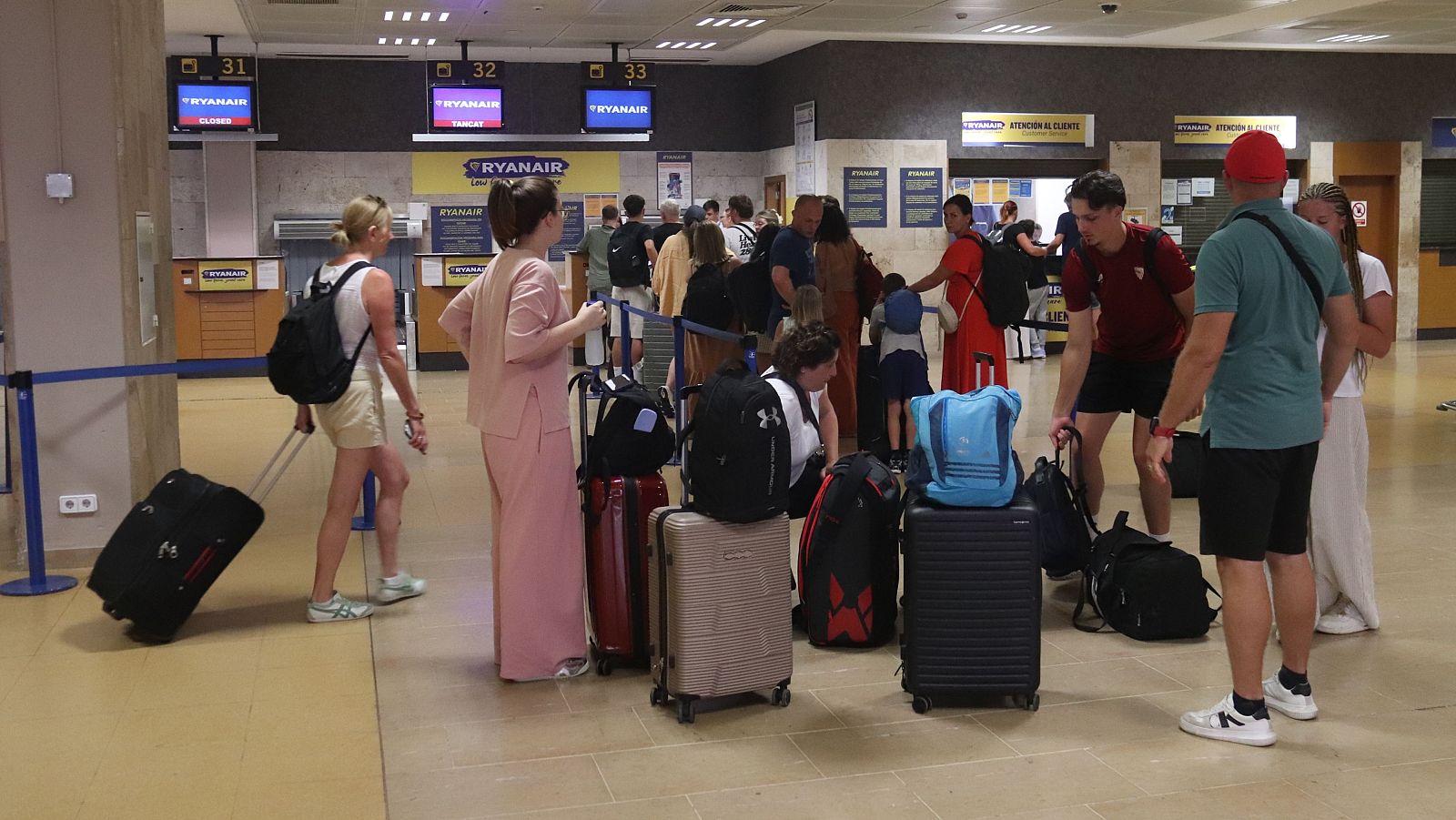 Pasajeros esperan en cola para facturar en mostradores de Ryanair (números 31-33 visibles).  Maletas y mochilas presentes.