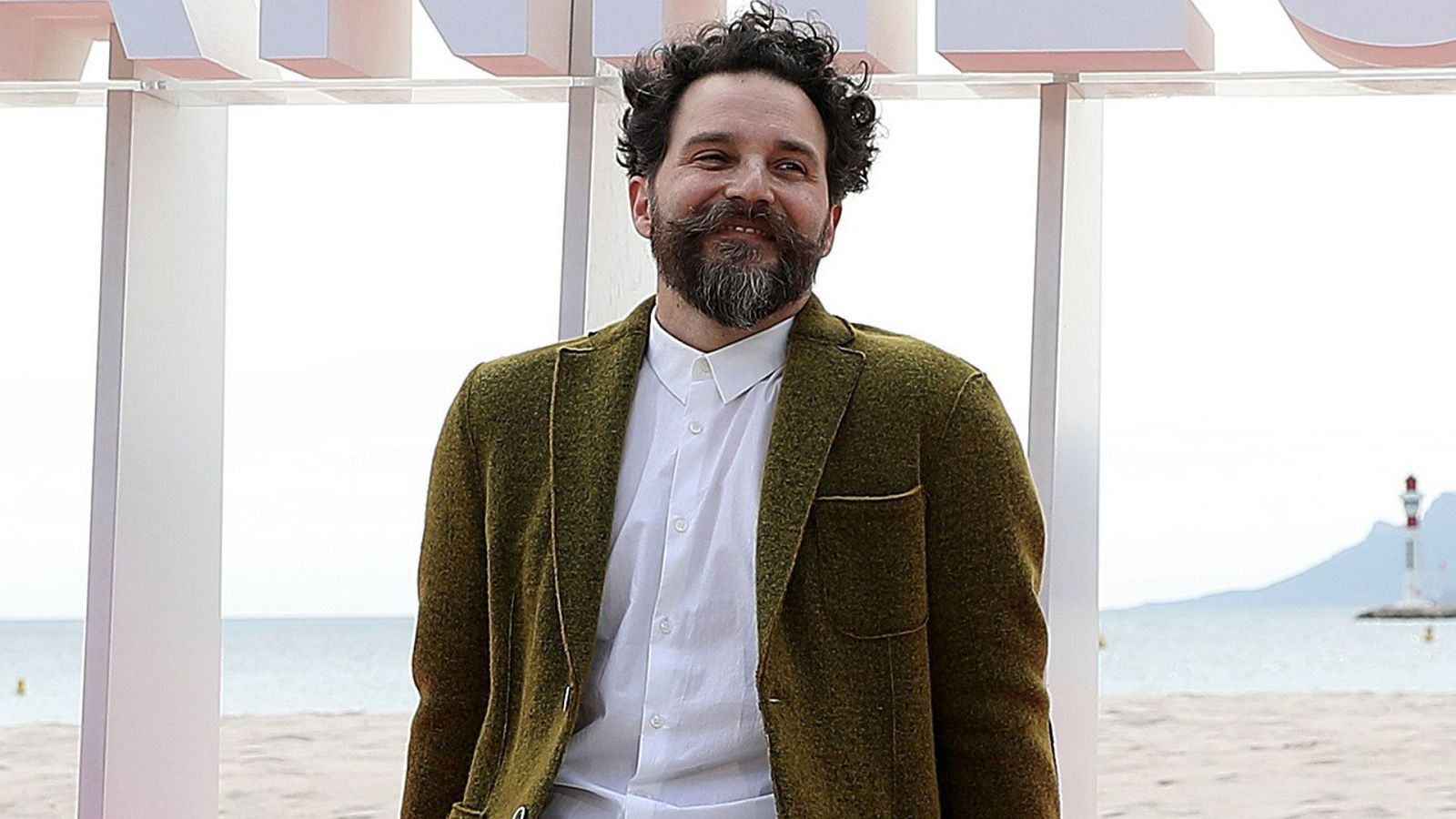 Compositor con chaqueta verde oliva y camisa blanca, frente a un fondo de playa y faro.  Texto: "Mañana más".