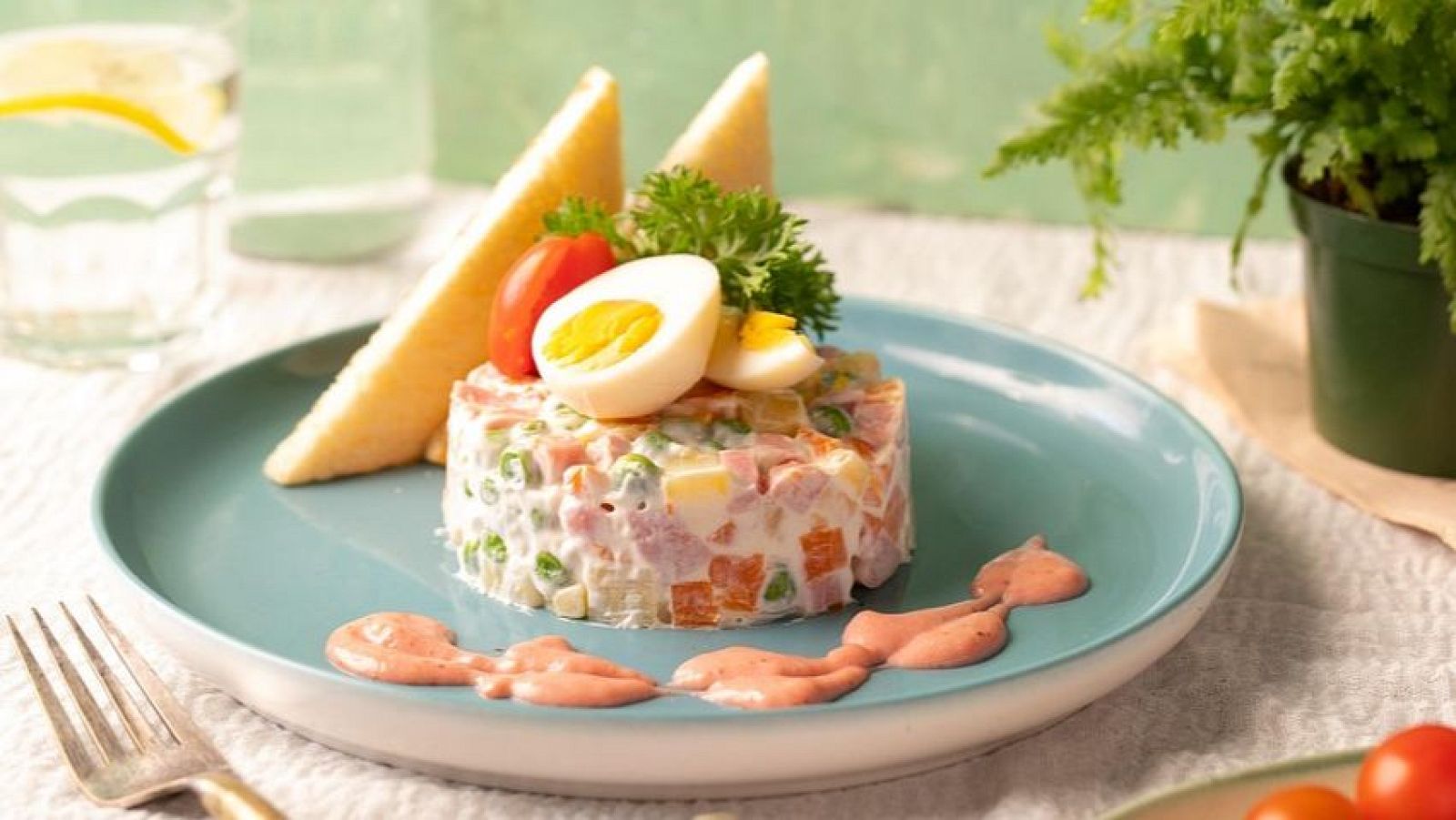 Ensalada rusa cilíndrica con guisantes, zanahoria, patata, huevo y embutido en mayonesa, decorada con pan tostado, perejil y tomate cherry, servida en plato azul verdoso.