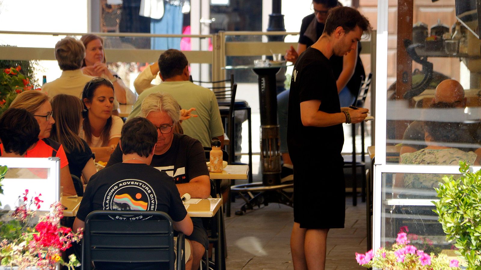 Terraza de restaurante con clientes sentados en mesas, un camarero toma nota y plantas decorativas. Ambiente relajado.