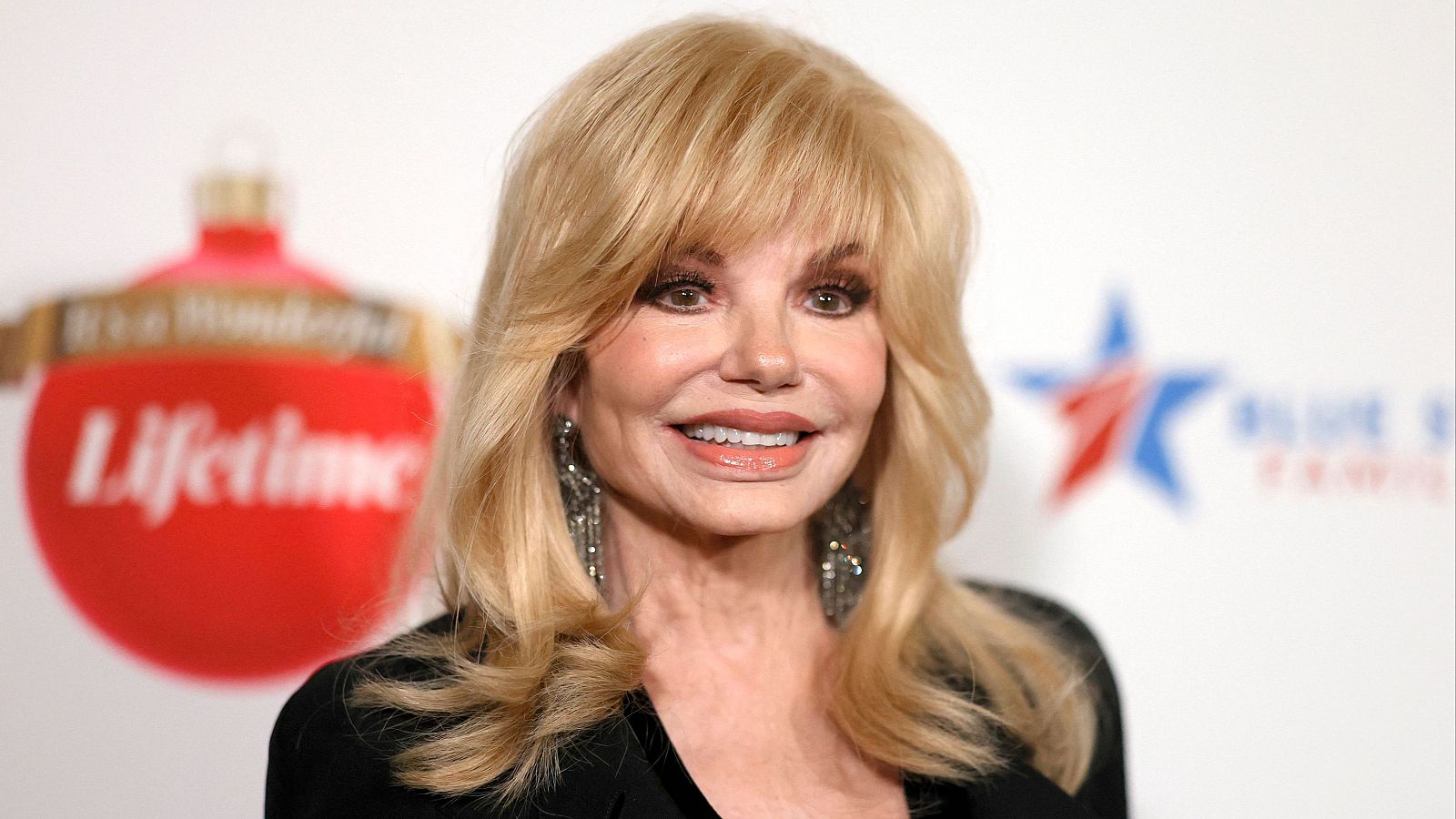 Retrato sonriente de Loni Anderson en 2003 con cabello rubio, pendientes largos y un adorno navideño rojo con la palabra 'Lifetime' al fondo.