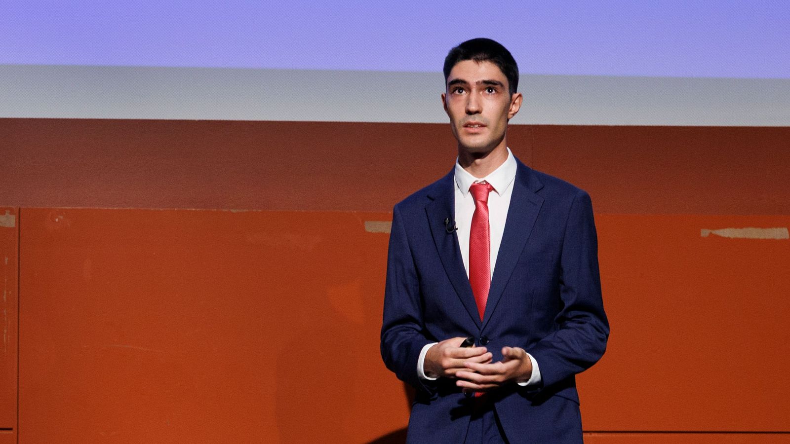 Presentación de un Trabajo Fin de Máster: un hombre en traje azul marino y corbata roja expone ante una pared con franjas horizontales, durante una jornada sobre discursos de odio.