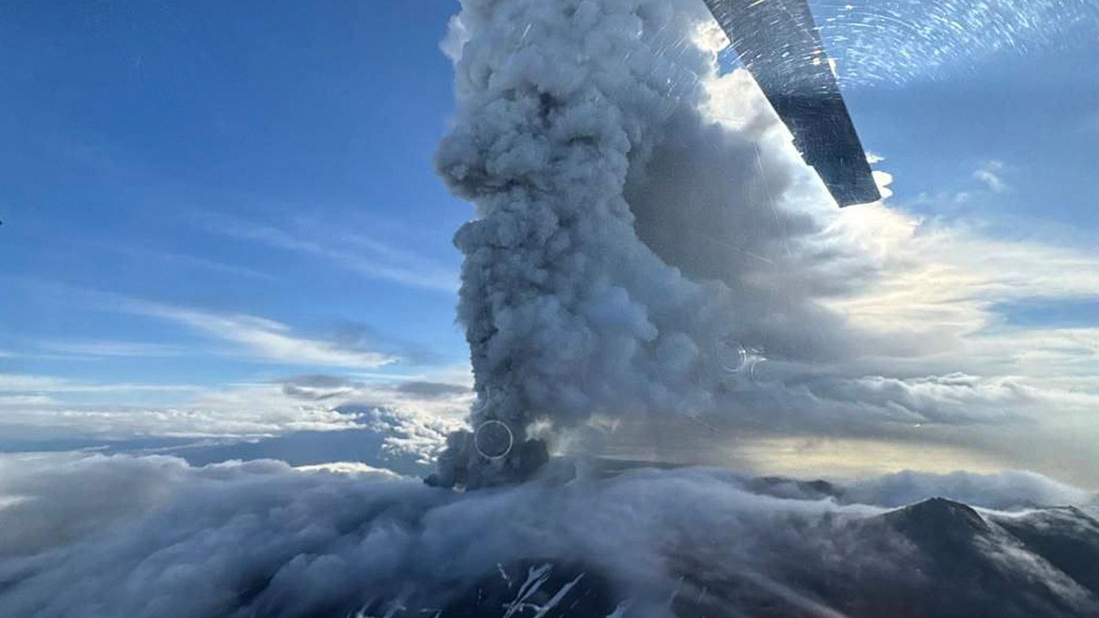 Erupción volcánica vista desde el aire: columna de humo y ceniza gris oscuro que se eleva sobre un paisaje montañoso con zonas nevadas.