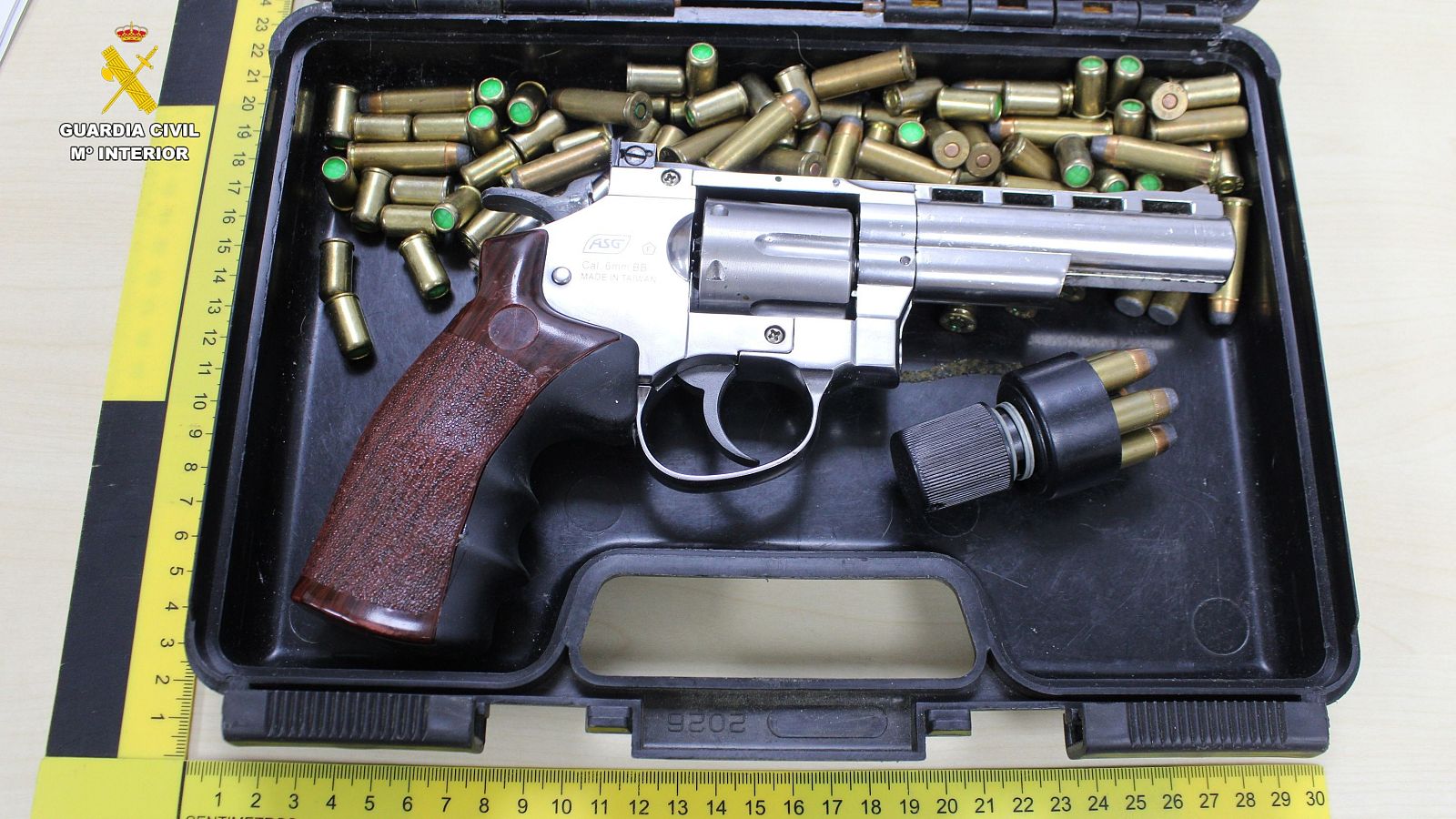 Revólver con empuñadura de madera, munición (puntas verdes) y cargador rápido en caja negra con regla métrica.  Logo Guardia Civil visible.