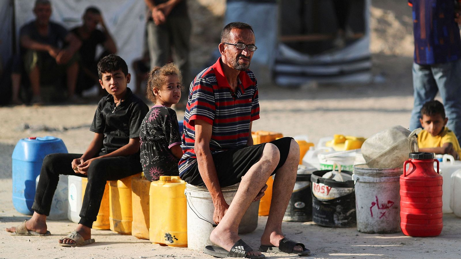 Personas, mayormente niños, sentados en cubos y bidones en un área polvorienta, esperando o recogiendo suministros.  Escena que refleja la escasez de recursos.
