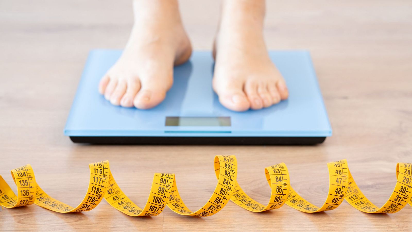 Pies sobre báscula azul en suelo de madera; cinta métrica amarilla desenfocada en primer plano.  Posible control de peso.