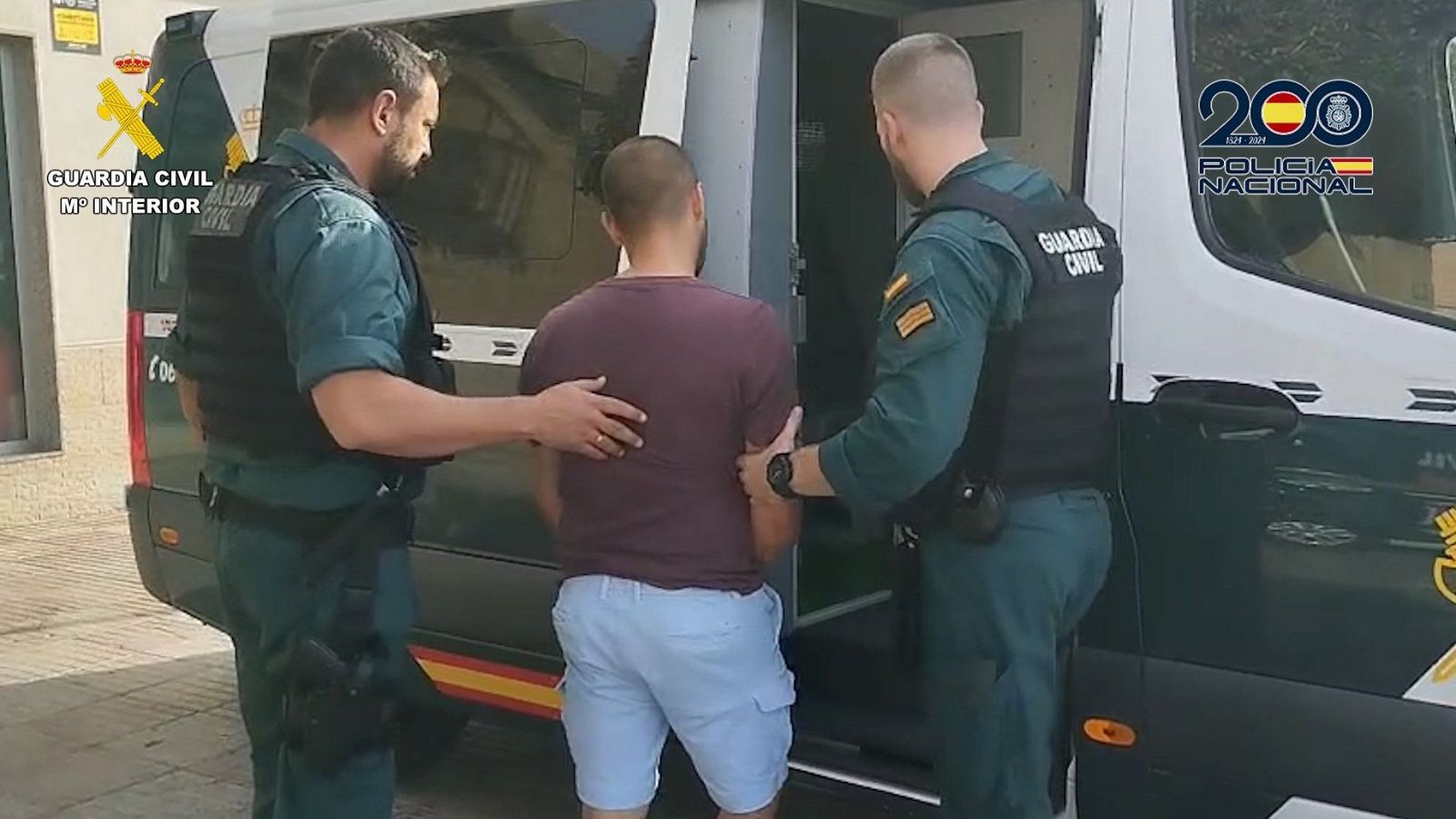 Detención de un hombre por dos agentes de la Guardia Civil. El detenido viste camiseta granate y pantalón corto azul. El vehículo policial muestra el logo del 200 aniversario de la Policía Nacional.