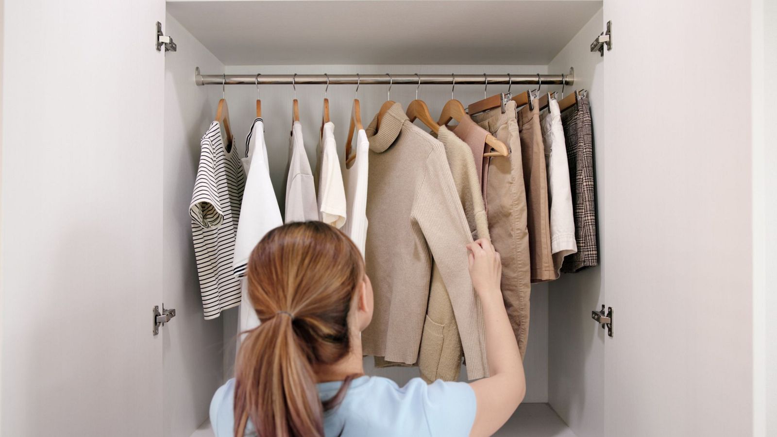 Mujer frente a armario blanco con ropa colgada en perchas de madera. Prendas en tonos neutros, ordenadas.  Posible selección de vestuario.