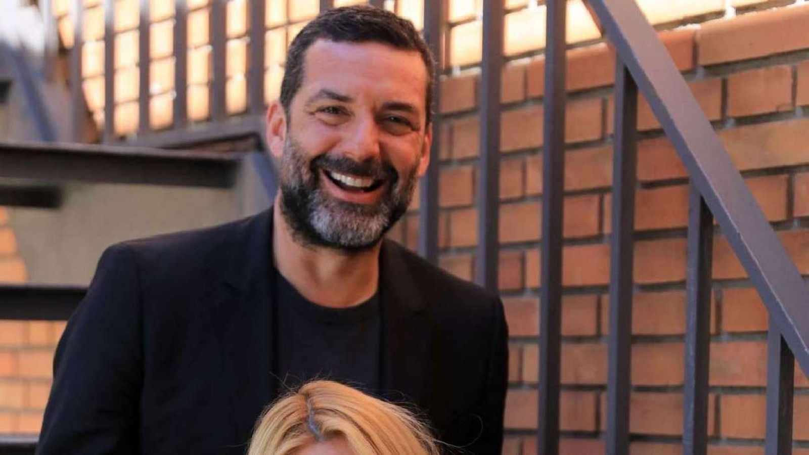 Jaume Anglada sonriendo, pelo corto y oscuro, barba canosa, americana negra sobre camiseta negra. Fondo: pared de ladrillo y barandilla metálica.