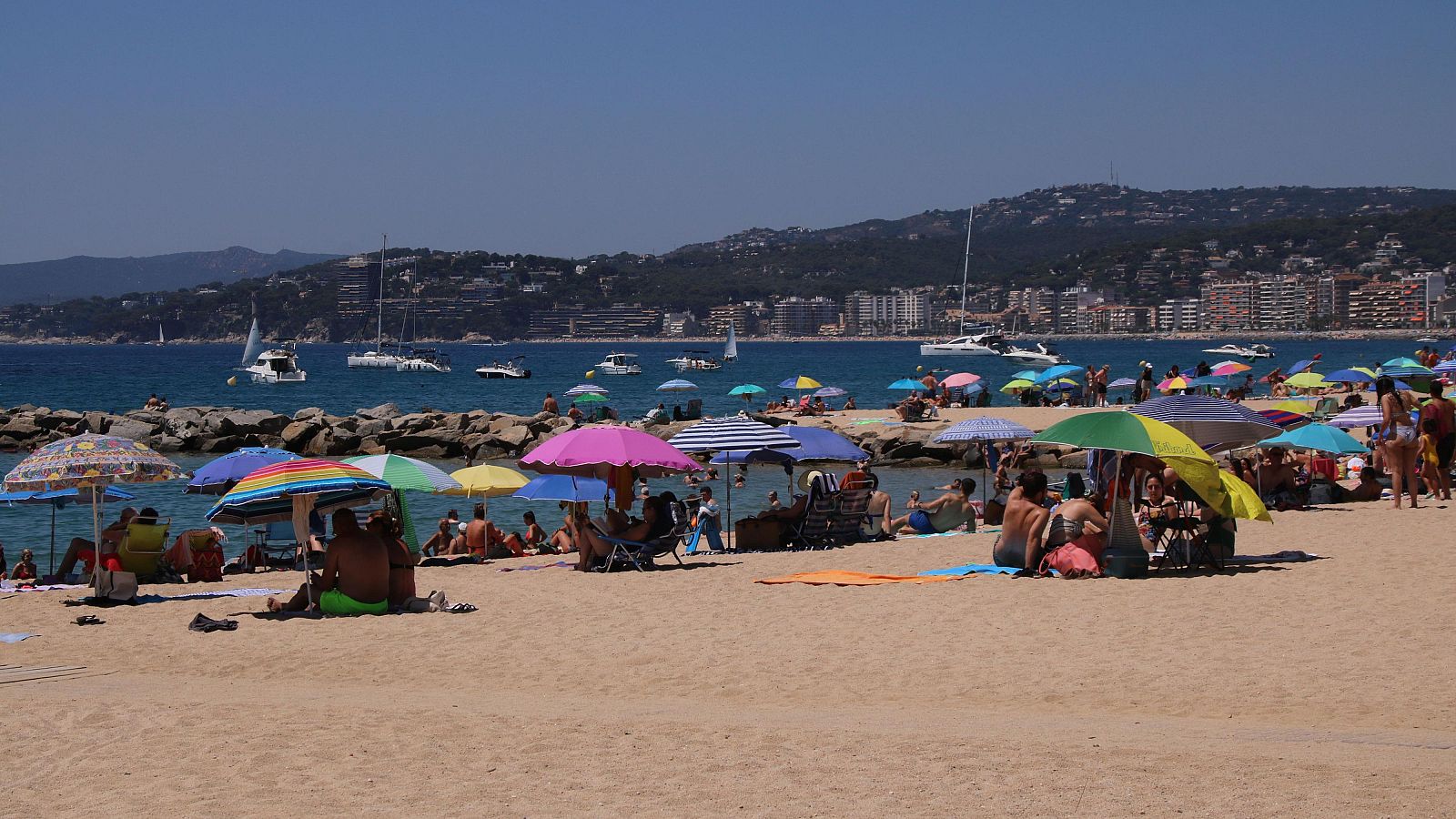 Playa abarrotada en día soleado; gente en la arena, bajo sombrillas, bañándose y embarcaciones en el mar.  Ambiente veraniego.