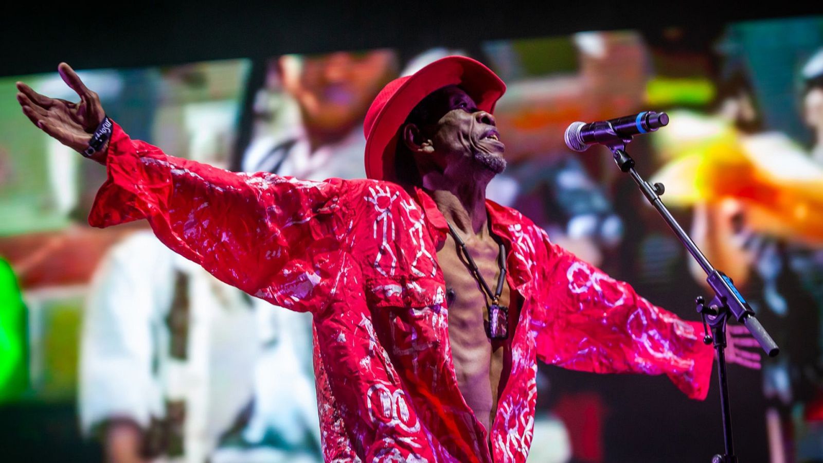 Hombre con sombrero y camisa roja actuando en un escenario con micrófono, fondo borroso y colores vibrantes.
