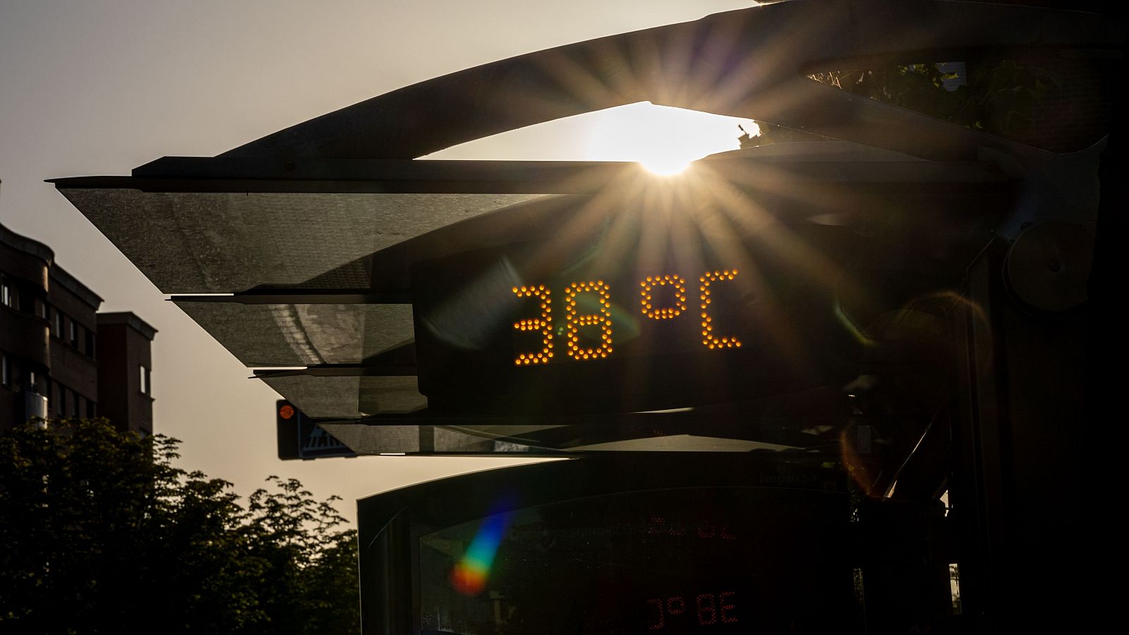 Termómetro digital naranja indica 38°C bajo un sol brillante.  Fondo urbano parcialmente visible.