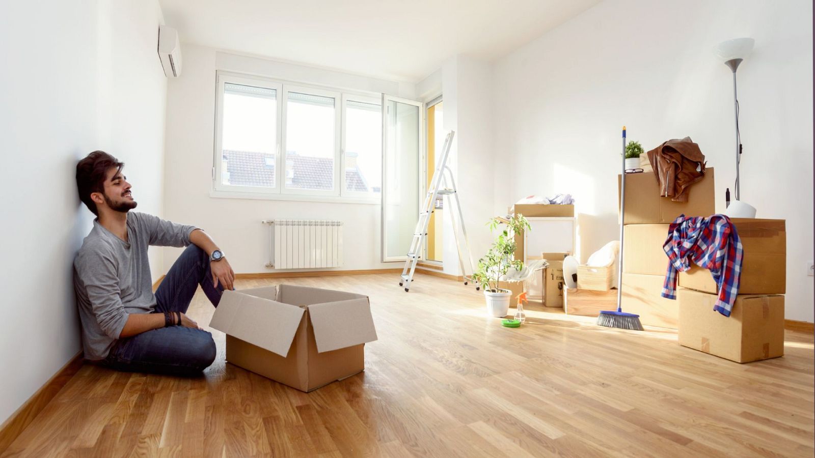 Joven relajado sentado en suelo de piso con cajas de cartón, escalera y otros enseres.  Ambiente luminoso y espacioso, sensación de mudanza y nuevo comienzo.
