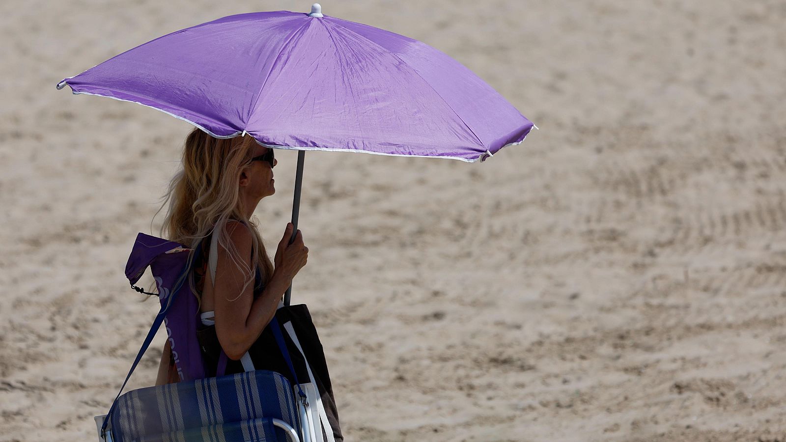 Mujer en la playa con sombrilla morada, gafas de sol y bikini, protegiéndose del sol con una silla plegable y una bolsa de playa.