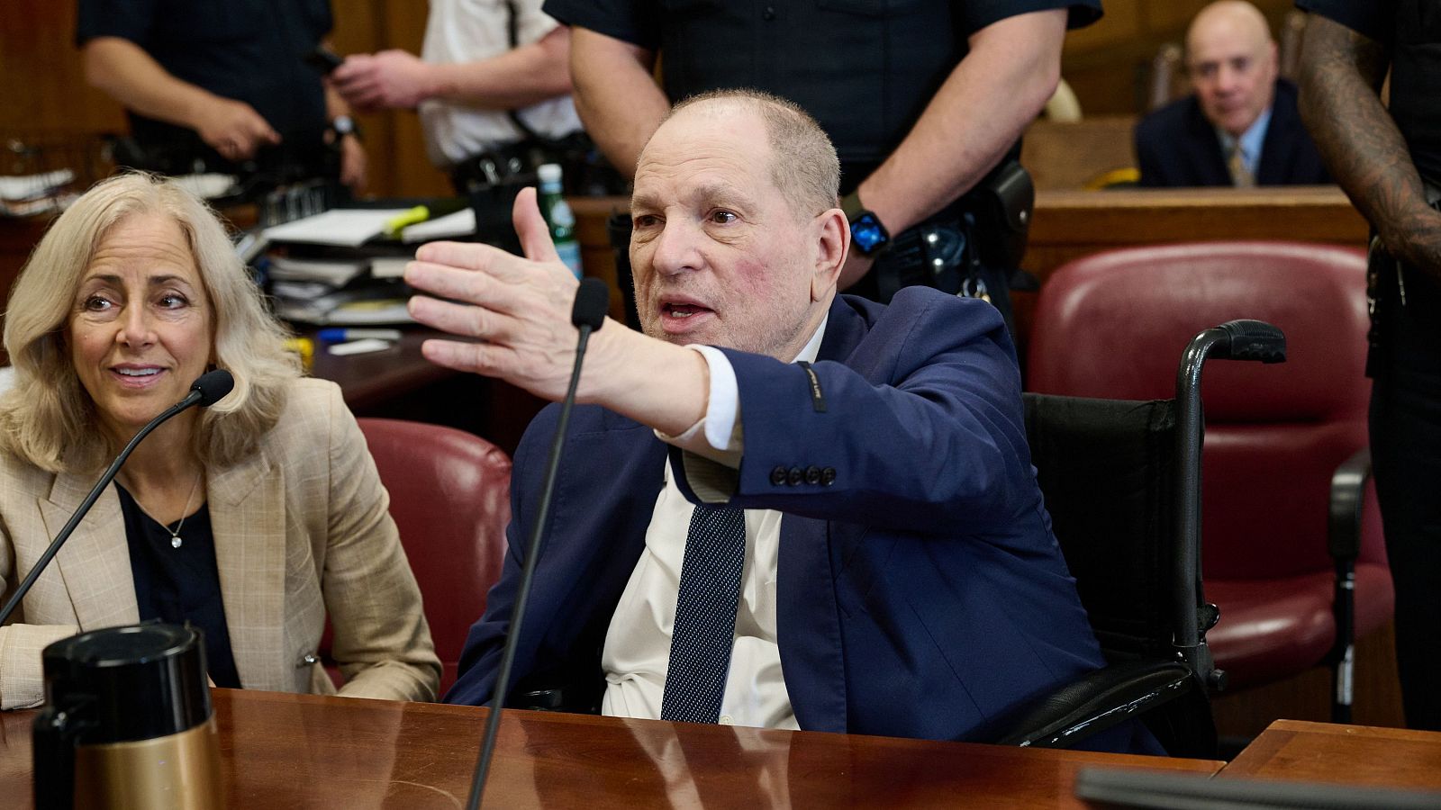 Imagen de Harvey Weinstein en silla de ruedas, traje oscuro y gesto con la mano derecha durante lo que parece ser una vista judicial.
