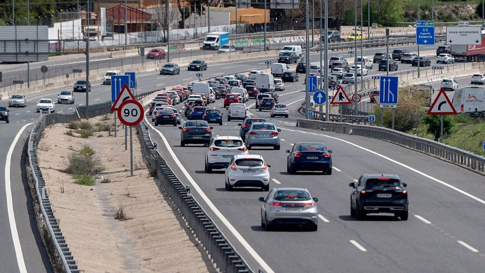 Autopista con tráfico intenso; varios vehículos circulan por múltiples carriles, con señales de velocidad y curvas visibles. Barrera metálica a un lado.