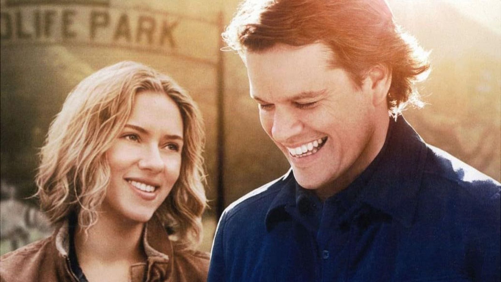 Scarlett Johansson, con chaqueta marrón, y Matt Damon, con camisa azul, sonríen mutuamente en una escena con un cartel desgastado que dice "OLIFE PARK" al fondo.