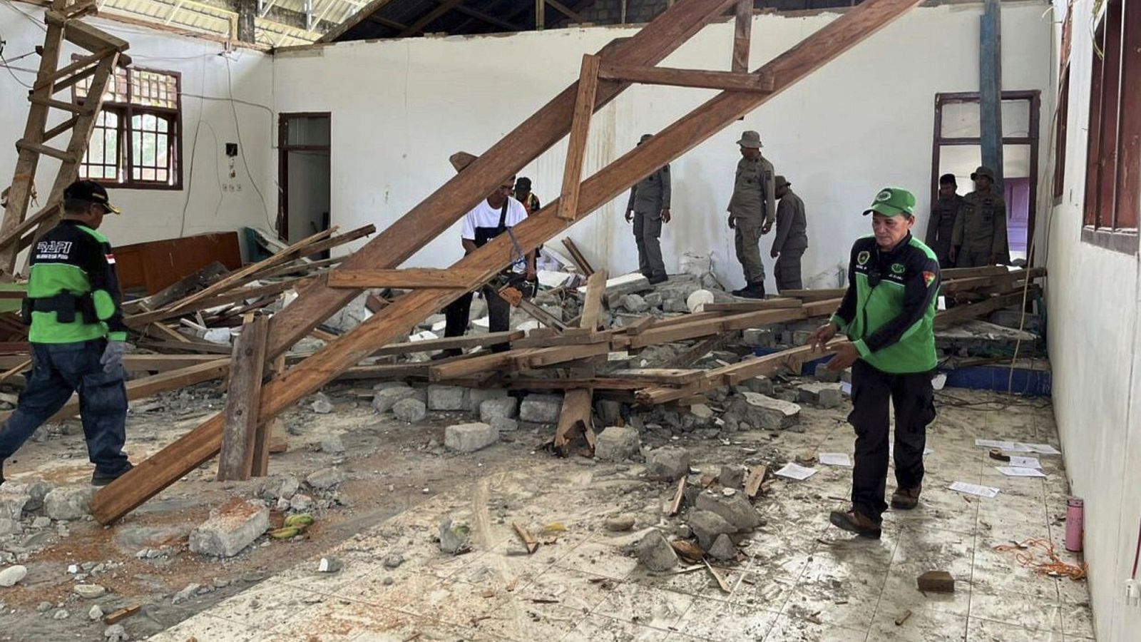 Edificio dañado por terremoto: Rescatistas, algunos con uniformes verdes, trabajan con motosierras entre escombros de madera y materiales de construcción.  Otros evalúan los daños.