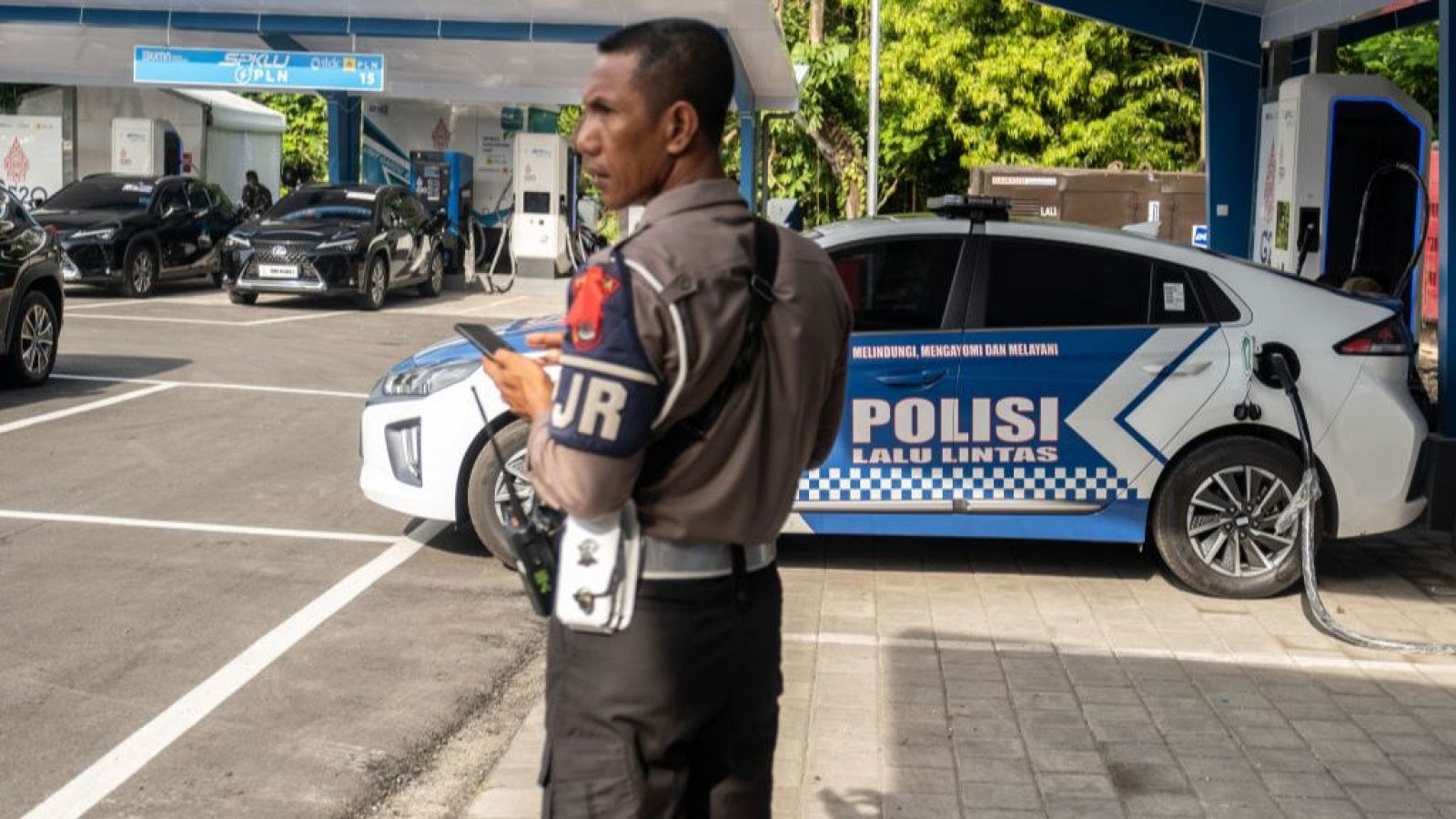 Policía indonesio con brazalete 'JR' mira su móvil junto a un coche eléctrico policial en una estación de carga.  Otros vehículos, algunos eléctricos, están aparcados en la zona.