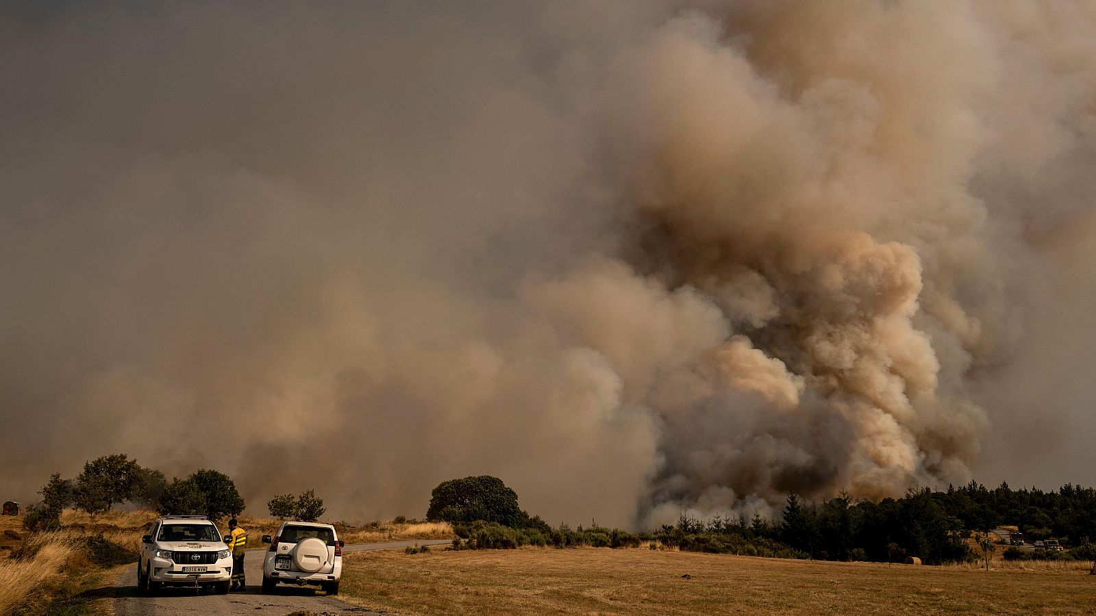 Incendio forestal de gran magnitud.  Dos vehículos y personal cerca del fuego, evaluando la situación en un terreno seco y arbolado.